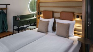 Una habitación de hotel moderna con una cama grande, un espejo redondo, una tocador con encimera verde y un cabecero de madera con iluminación integrada.
