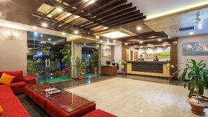 A hotel lobby with a reception desk, red seating area, and potted plants. - Hotel Queens Park (Pokhara)