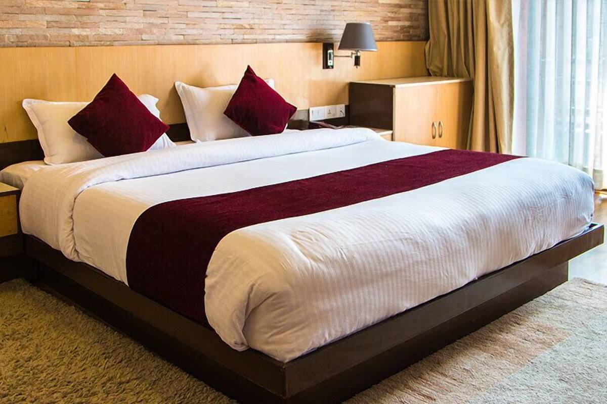 A large bed with white and red bedding, a wooden headboard, a nightstand with a lamp, and a view of the outdoors through a window.