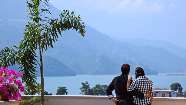 A couple standing on a balcony overlooking a lake with mountains in the background. - Hotel Queens Park (Pokhara)