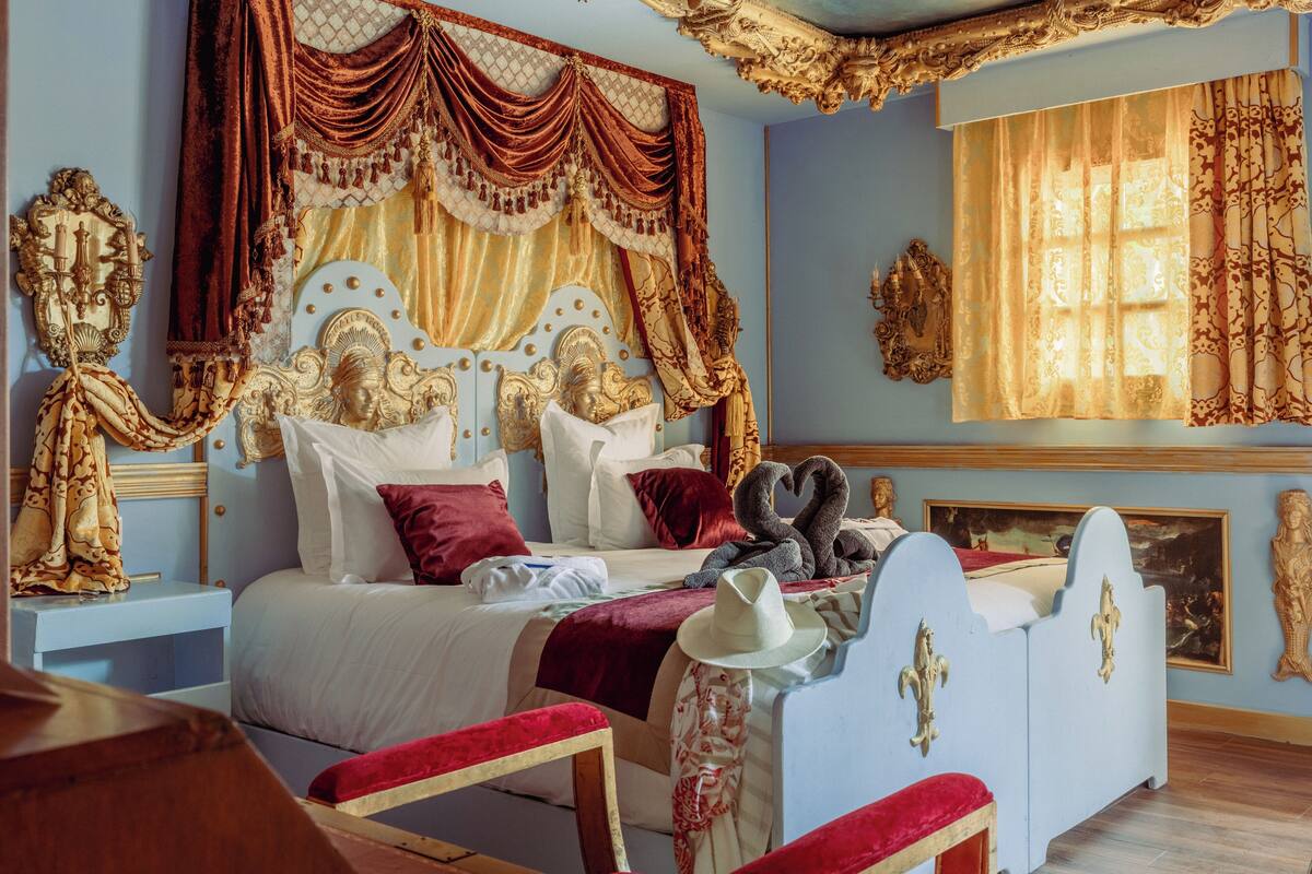 Un dormitorio con una cama grande, un cabecero ornamentado y un dosel. Se ve una mesita de noche con una lámpara y un espejo.