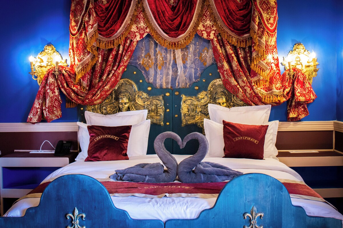 A luxurious bedroom with a bed featuring a heart-shaped towel arrangement, ornate drapery, and a blue headboard with fleur-de-lis motifs.