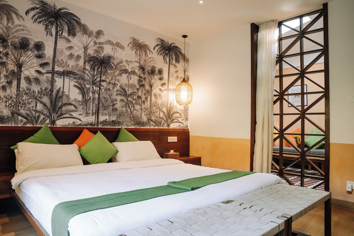 A hotel room with a large bed, a headboard with multiple pillows, a wall mural of palm trees, a hanging light fixture, and a view through a wooden lattice window.