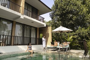 A pool area with a lounge chair, a sun umbrella, and a building with balconies. - Kasara Resort - Chitwan National Park (Bharatpur)