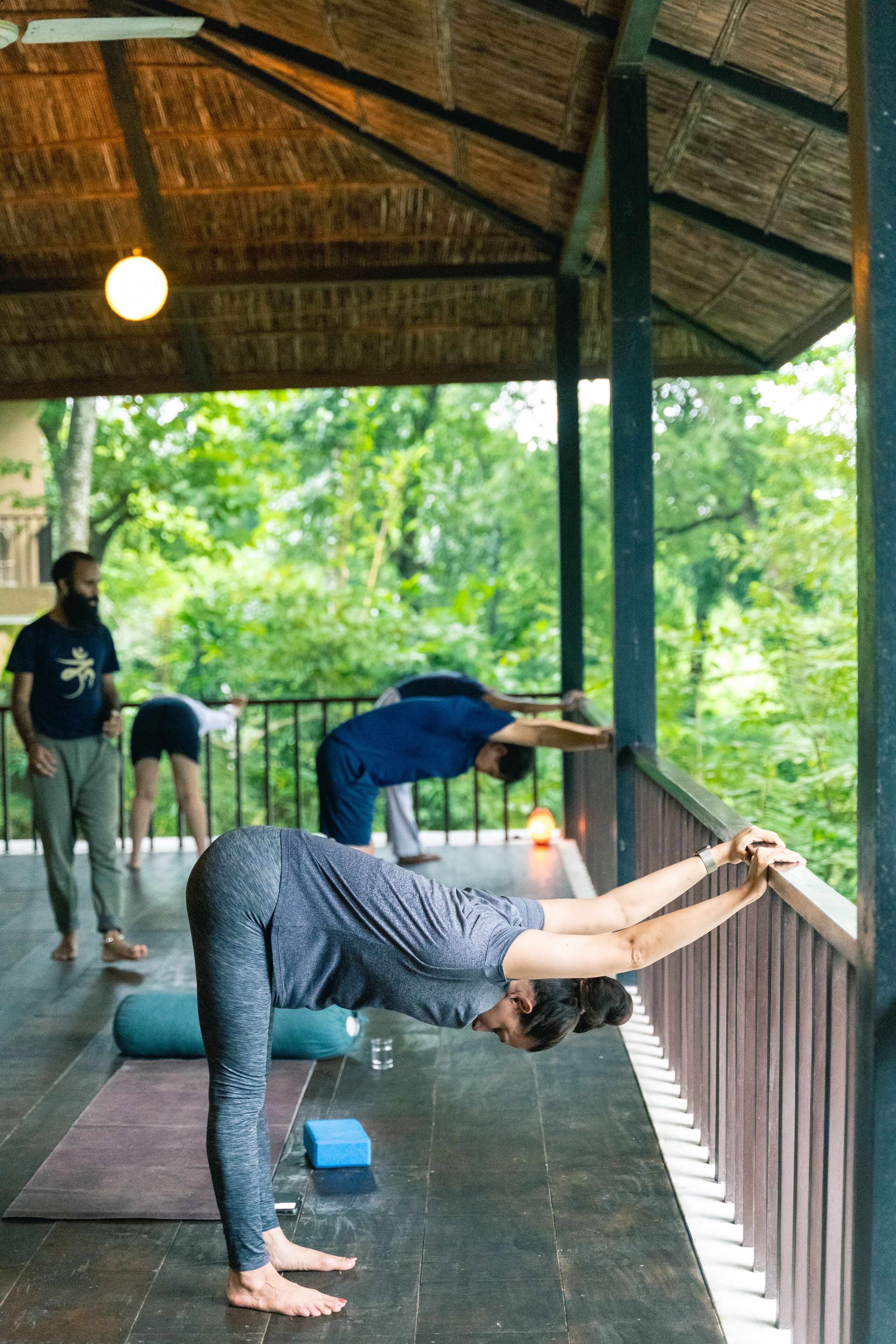 A yoga class in a covered outdoor area with wooden beams and a thatched roof.