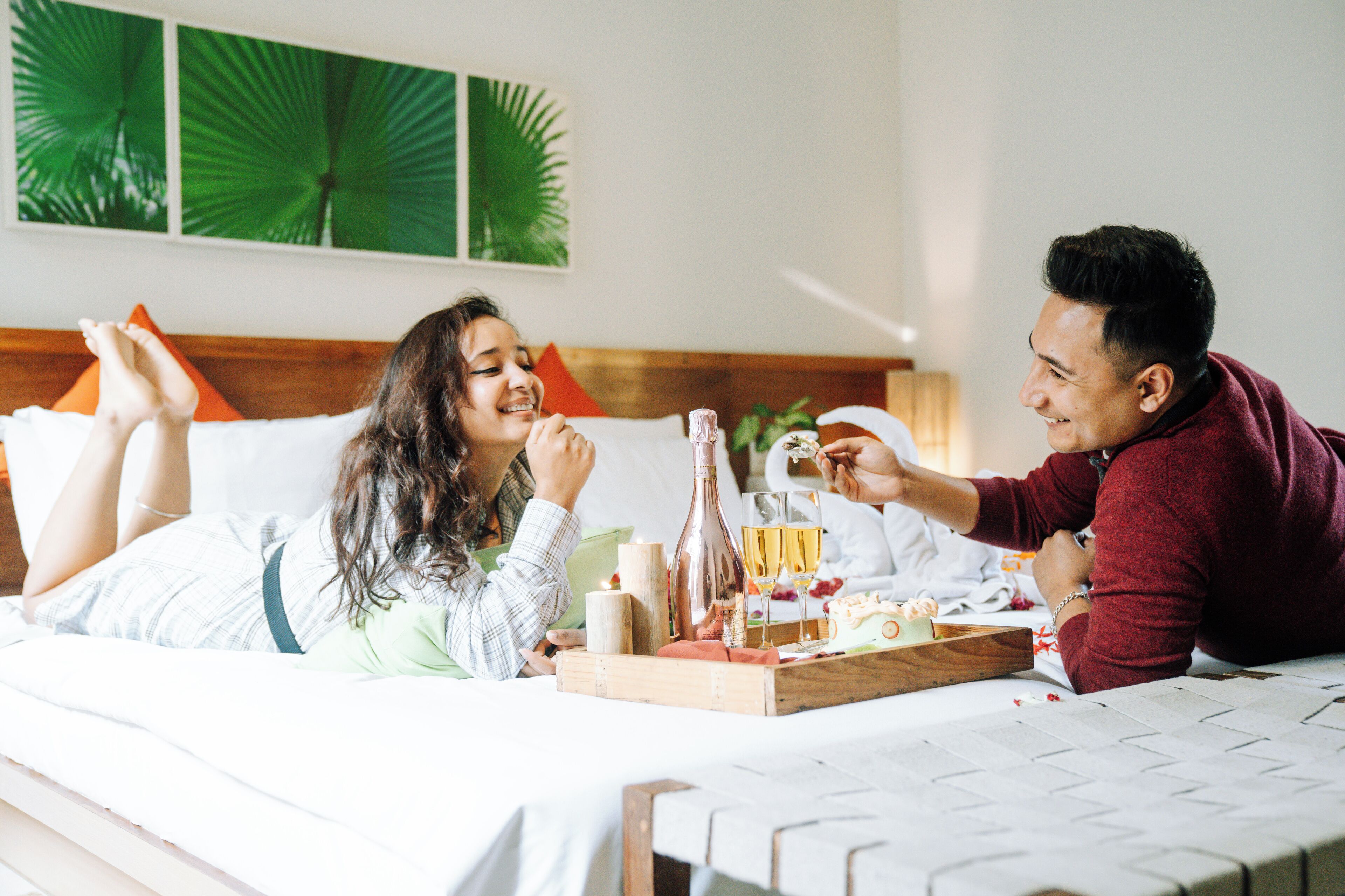 A couple lying on a bed with a tray of drinks and snacks.