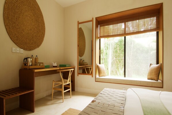 A bedroom with a wooden desk, a chair, a bed, a window with a view of greenery, and a large woven wall hanging.