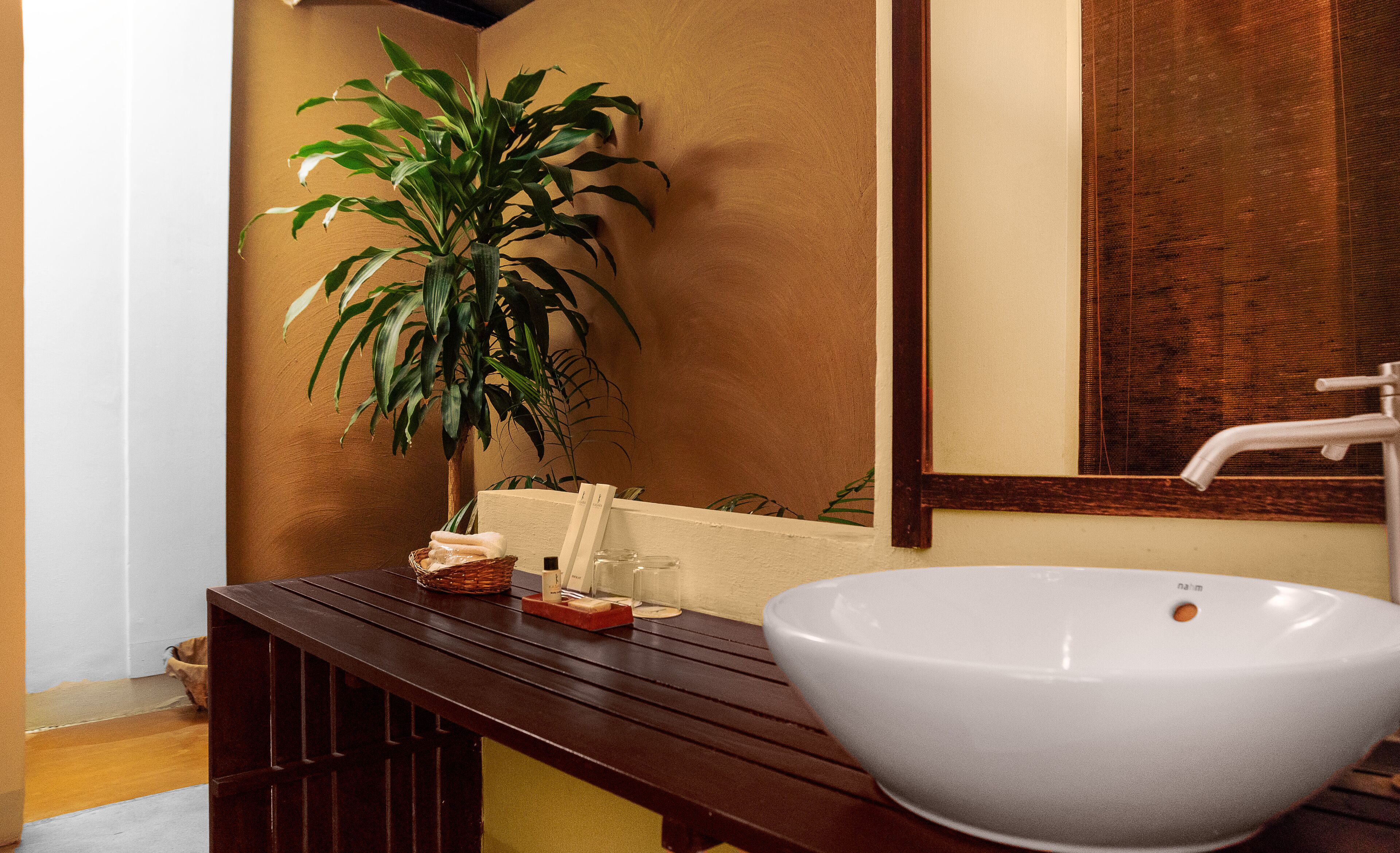 Un baño con un lavamanos grande de color blanco, un banco de madera, una planta en maceta y un espejo.