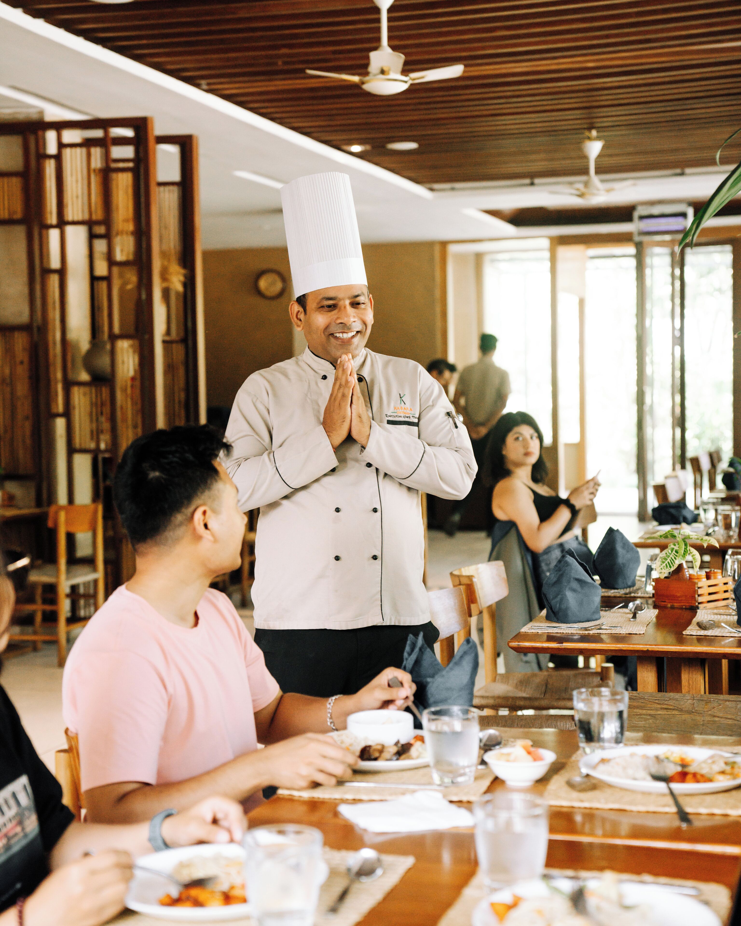 L’intérieur d’un restaurant, avec des meubles en bois, un ventilateur de plafond et une personne vêtue d’un uniforme de chef, debout, les mains jointes.