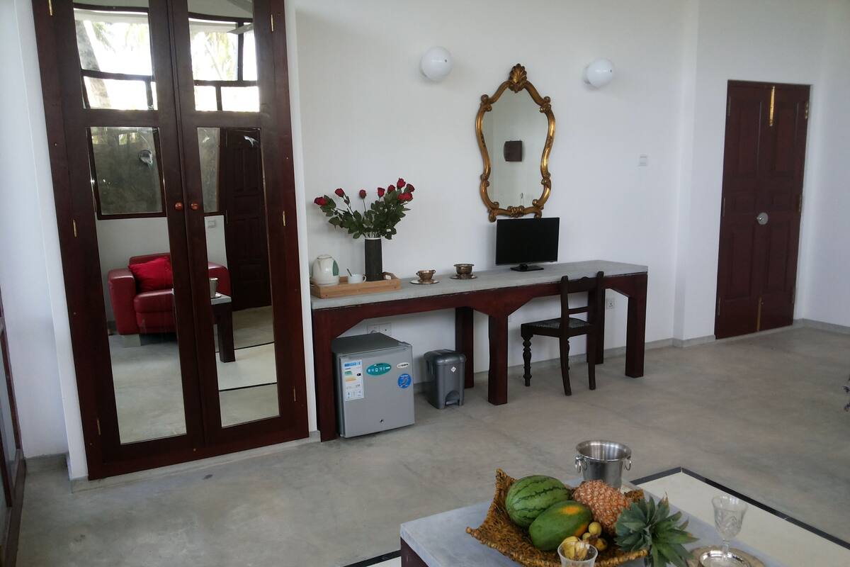 Une pièce avec un bureau, une chaise et un miroir au mur. Au fond, on aperçoit un canapé rouge, et sur le sol, une petite table avec un panier de fruits et un verre.