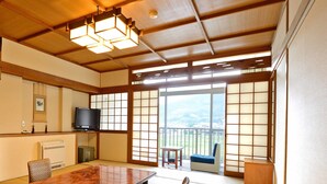 Ein traditioneller japanischer Raum mit Tatami-Böden, einem Holztisch, Tatami-Matten, einem Fernseher und Blick auf eine Berglandschaft durch große Schiebetüren.