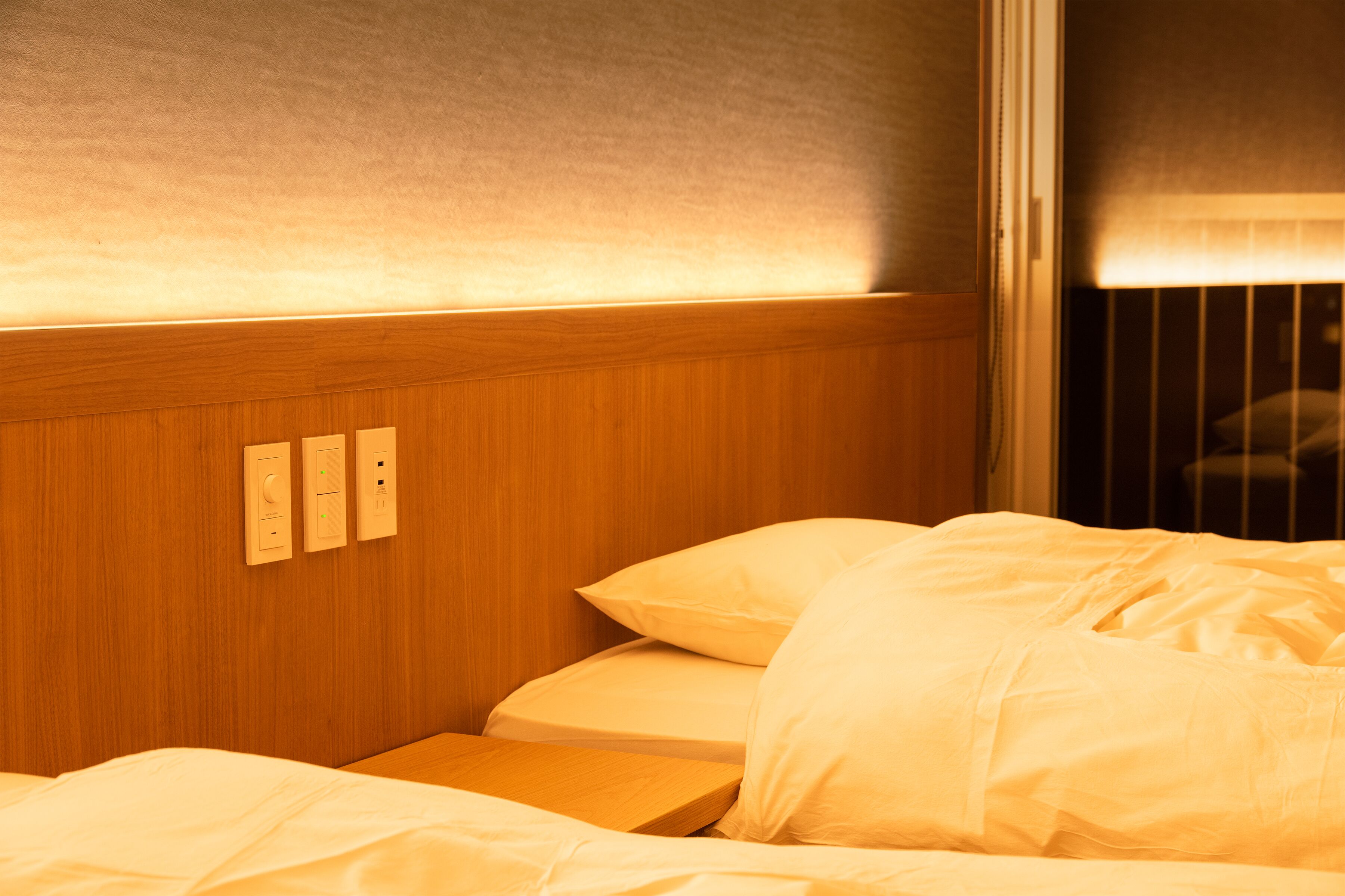 A neatly made bed with white linens, a wooden headboard with built-in lighting, and a window with blinds partially drawn.