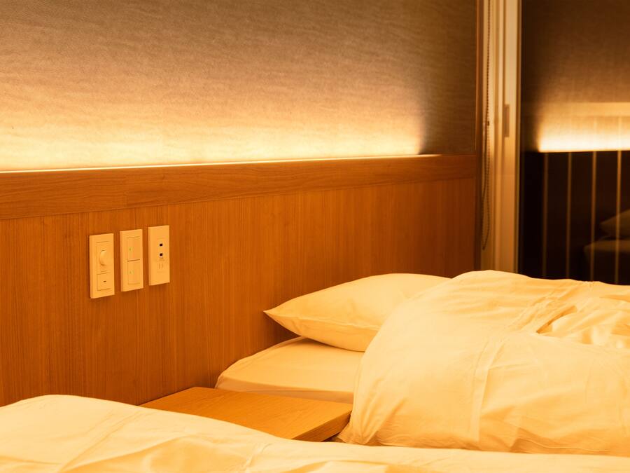 A neatly made bed with white linens, a wooden headboard with built-in lighting, and a window with blinds partially drawn.