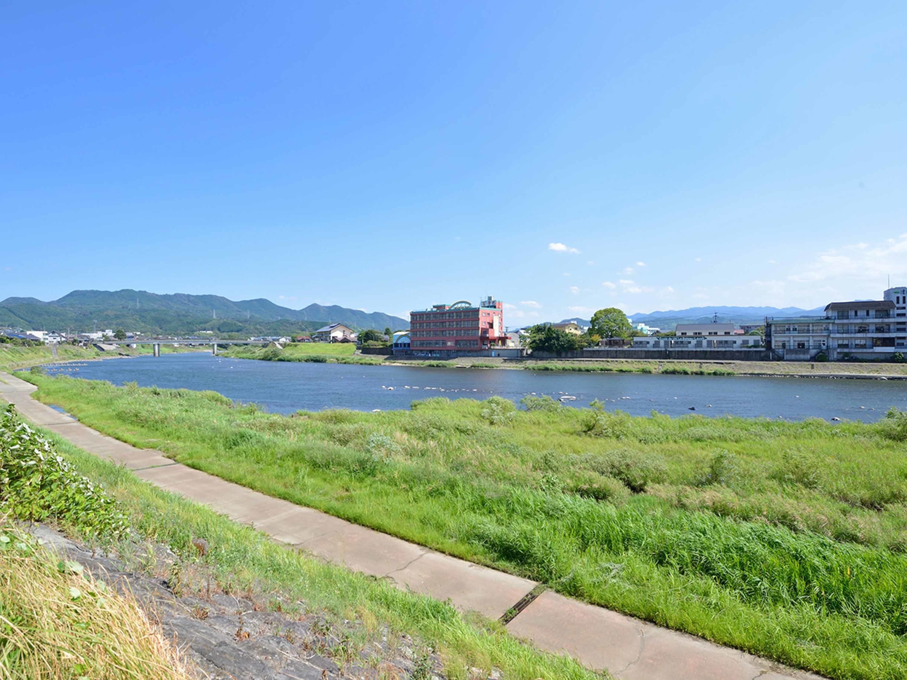 Un sendero junto al río con un camino de hormigón, un edificio rojo y un puente al fondo.