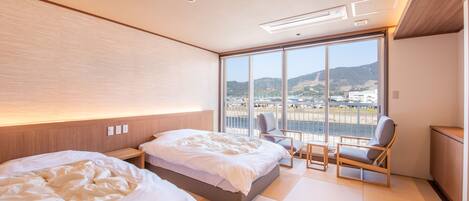 A modern hotel room with two beds, a large window with a view of a beach and hills, and a seating area with a chair and table.