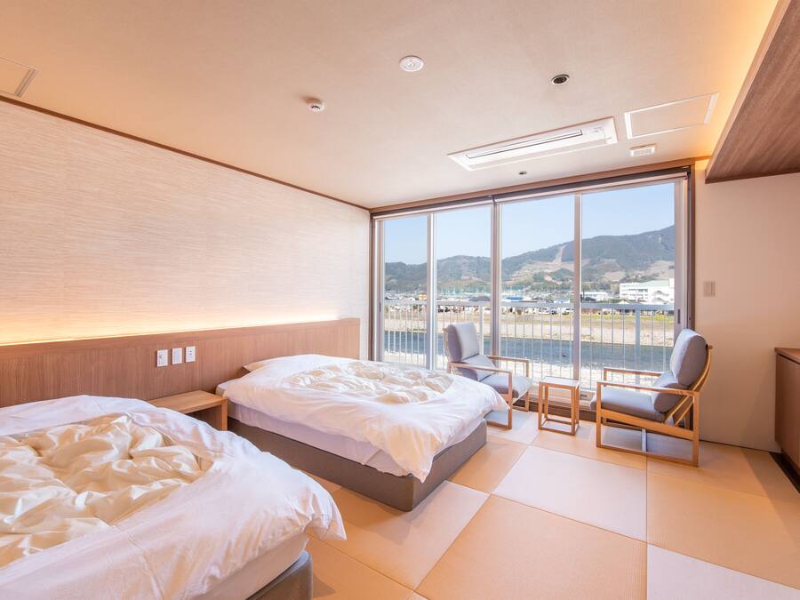 Une chambre d’hôtel moderne avec deux lits, une grande fenêtre donnant sur une plage et des collines, et un coin salon avec une chaise et une table.