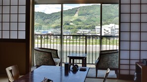 Ein traditioneller japanischer Raum mit Tatami-Böden, einem niedrigen Tisch und Stühlen. Ein großes Fenster bietet Blick auf einen grünen Hügel und ein Gebäude in der Ferne.