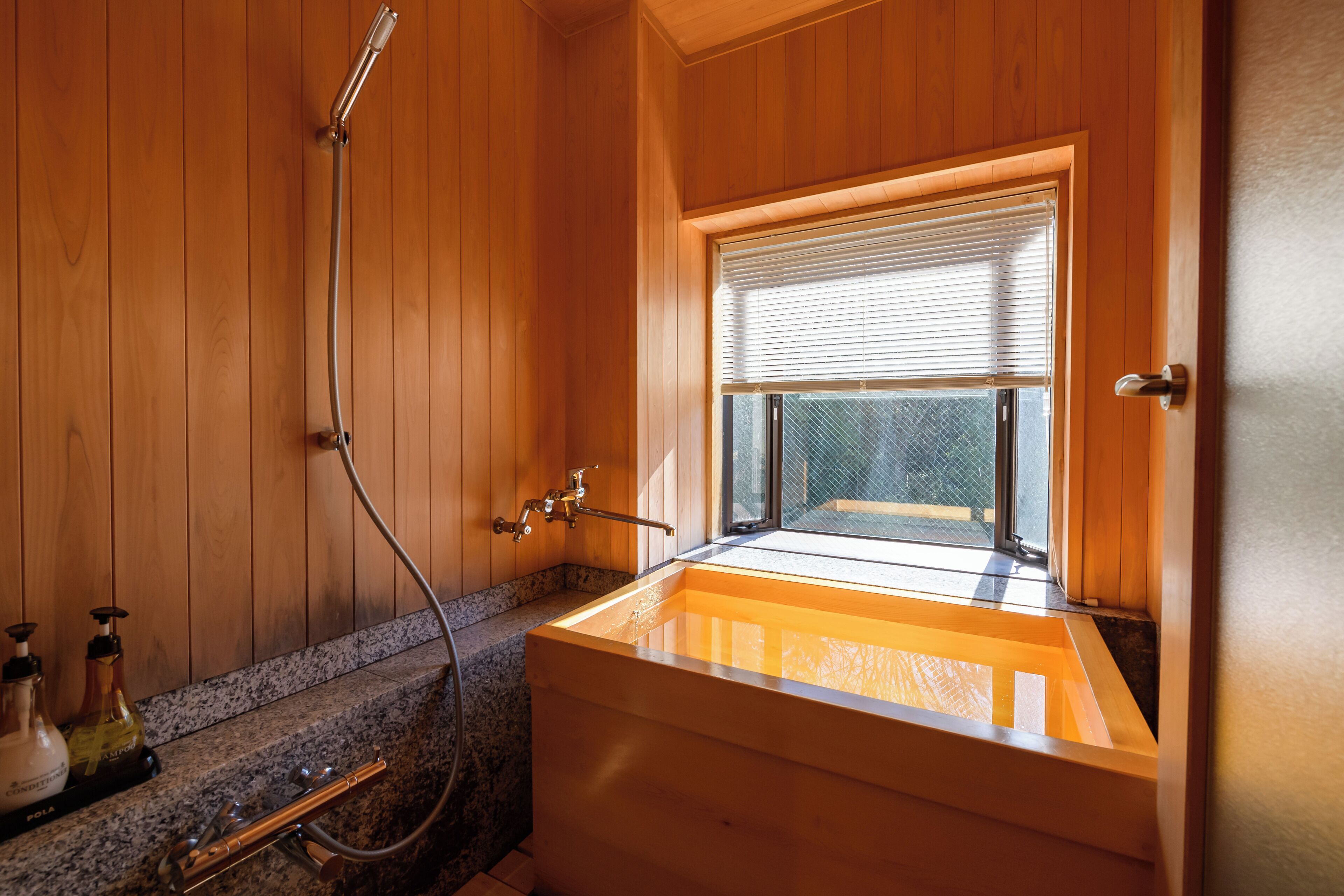 A wooden bathroom with a freestanding bathtub, a showerhead, and a window with blinds.