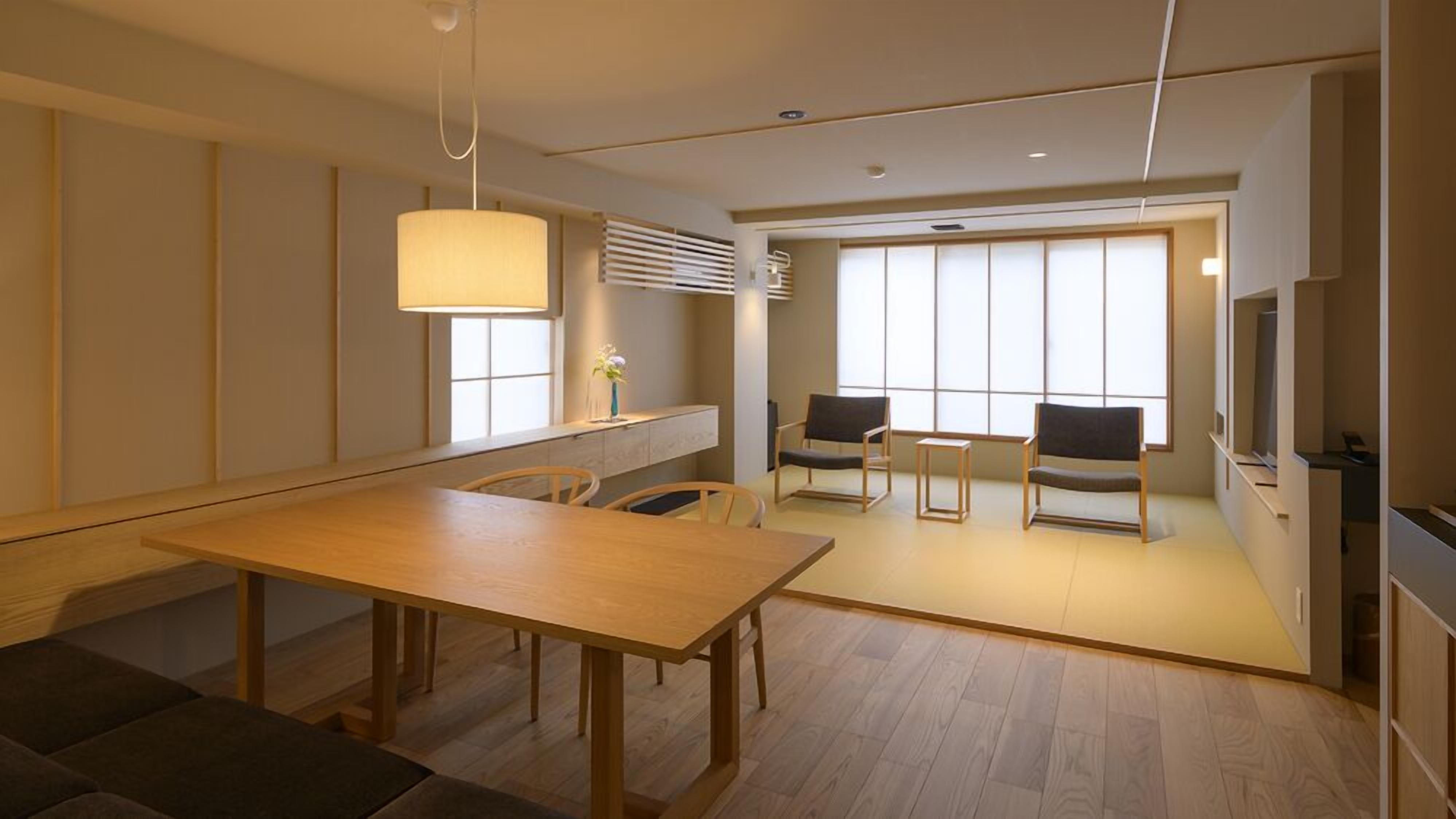 A modern hotel room with a wooden dining table, two chairs, a sofa, a TV, and a large window.