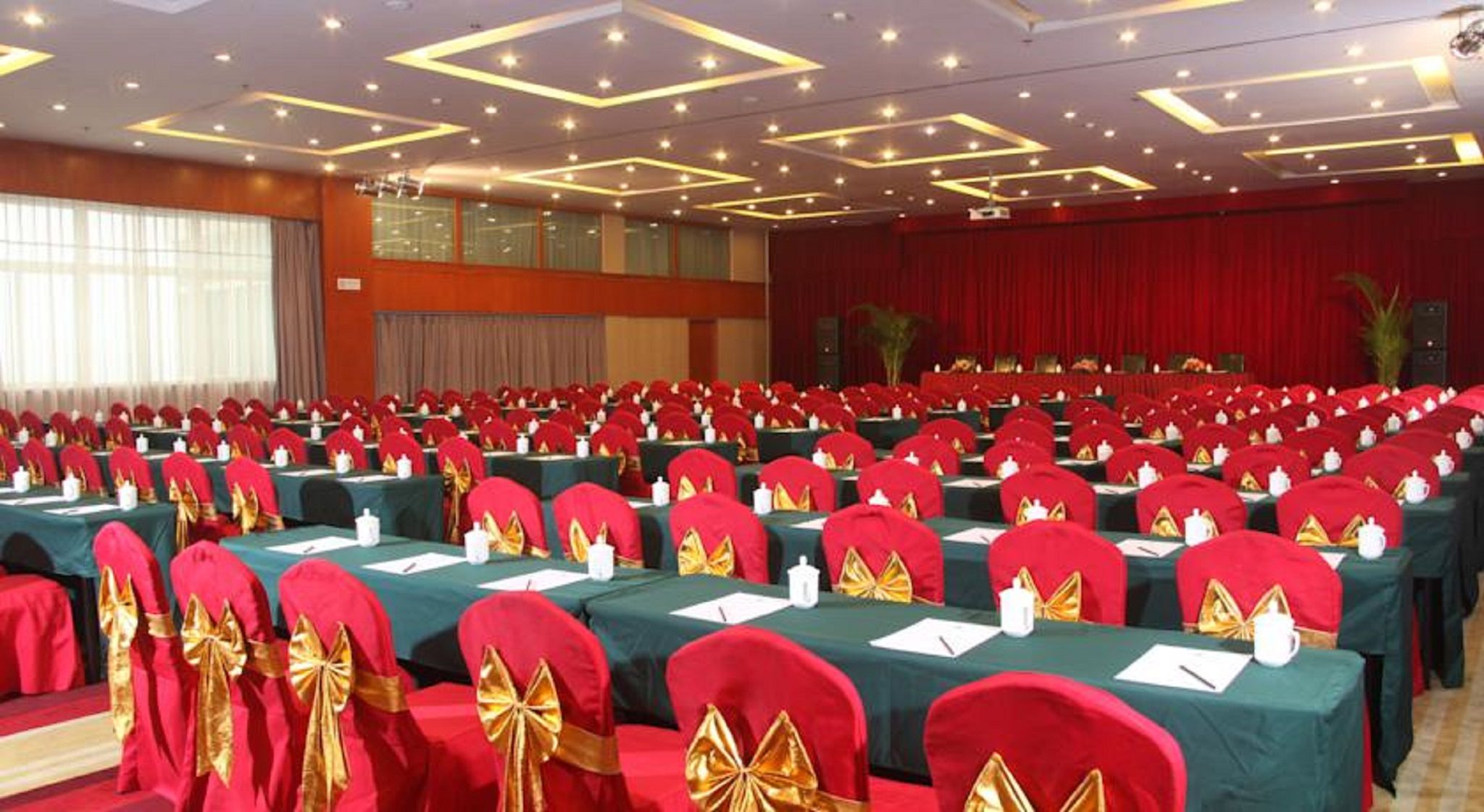 A conference room with rows of tables, red chairs with gold sashes, and a stage with a red curtain.