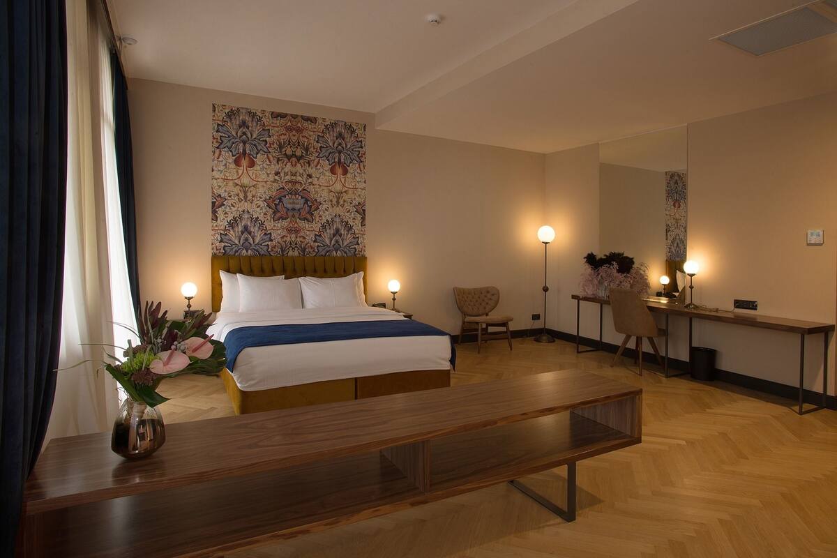 Una habitación de hotel con cama, escritorio, silla, un jarrón con flores y un espejo grande.