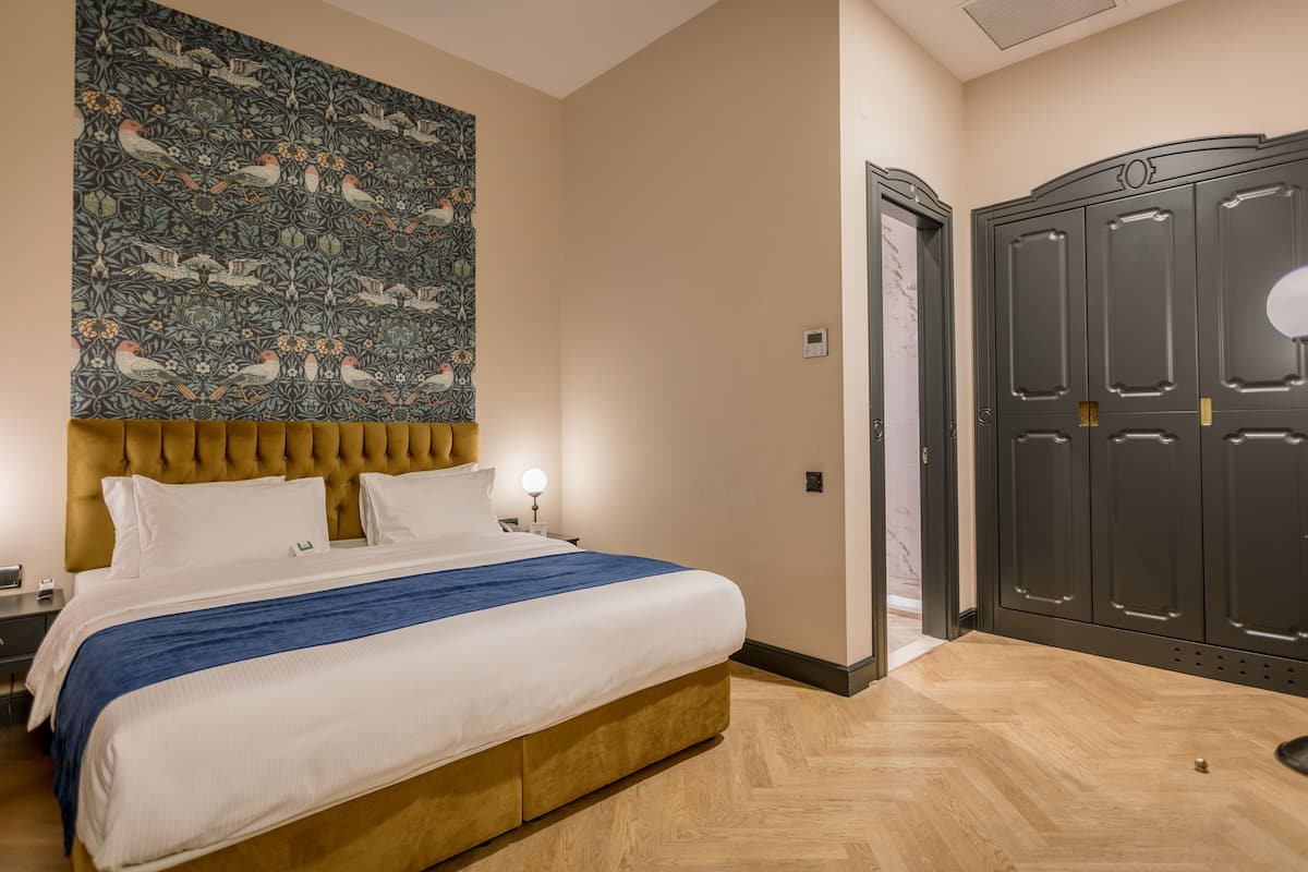 Habitación de hotel con una cama grande, cabecera con diseño capitonado, tapiz en la pared, armario y mesita de noche con lámpara.