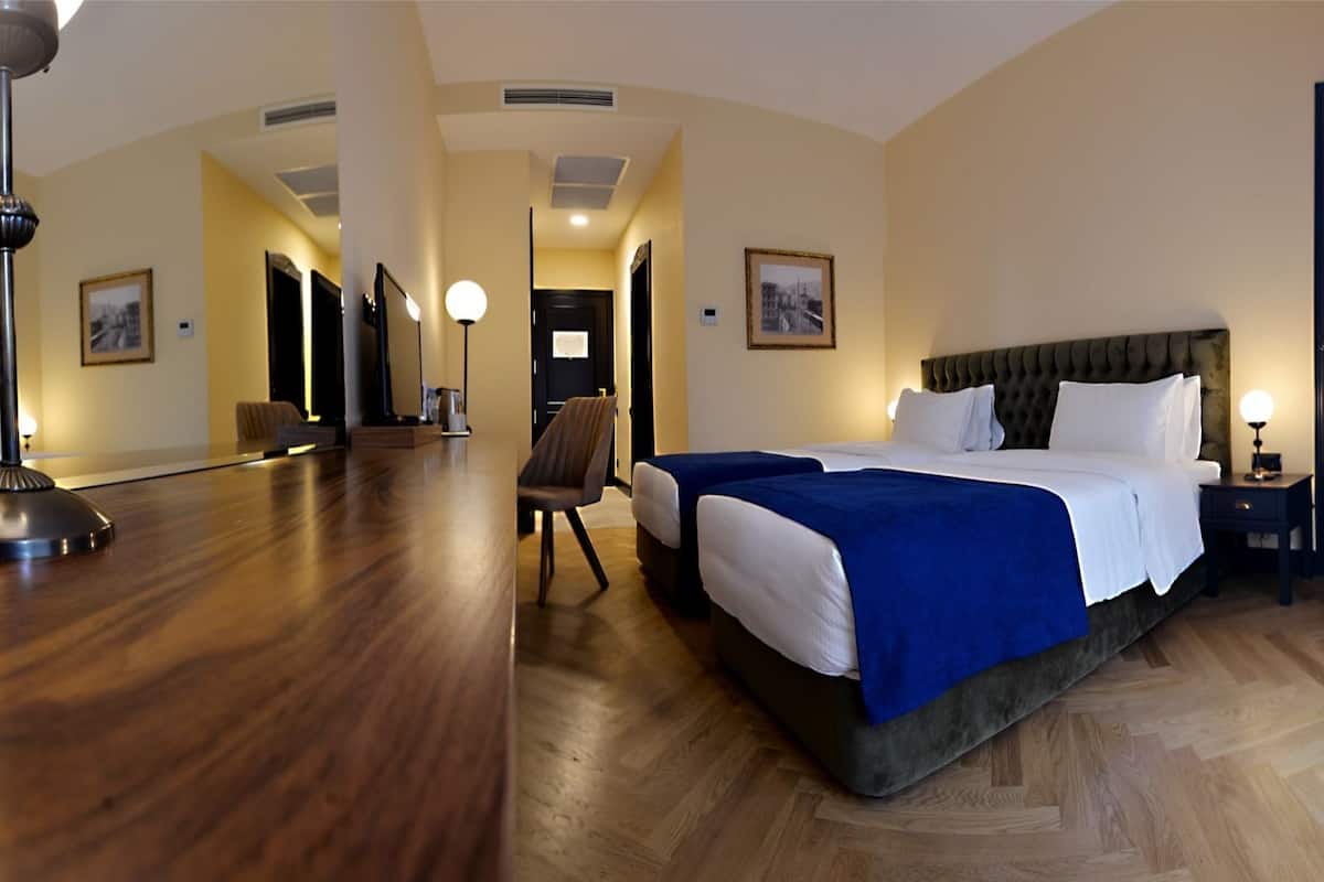 Una habitación de hotel con una cama grande, un escritorio y una silla. Hay dos cuadros en las paredes y una puerta a la derecha.