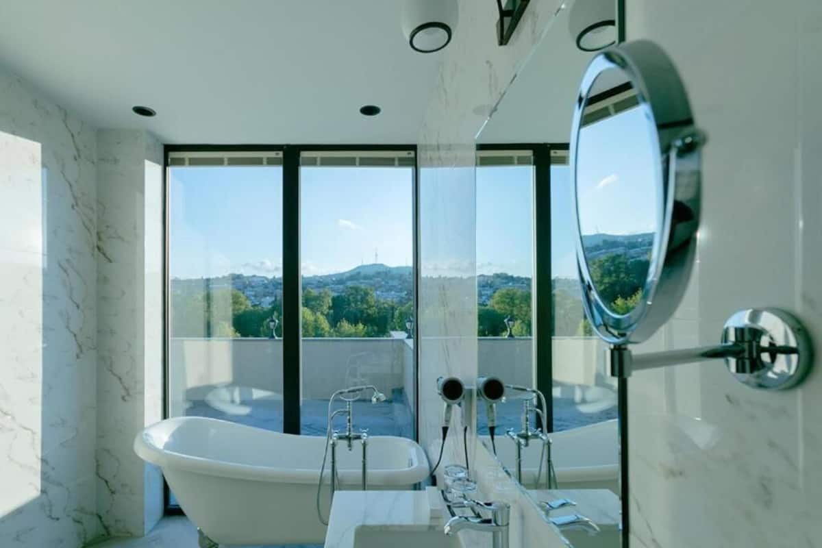 Un baño moderno con una bañera independiente, un espejo grande y vistas a un paisaje urbano a través de amplias ventanas.