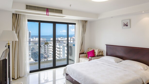 A hotel room with a large bed, a nightstand, a lamp, a window with curtains, and a view of the city. - Sanya Huabaoshi Hotel (Sanya)