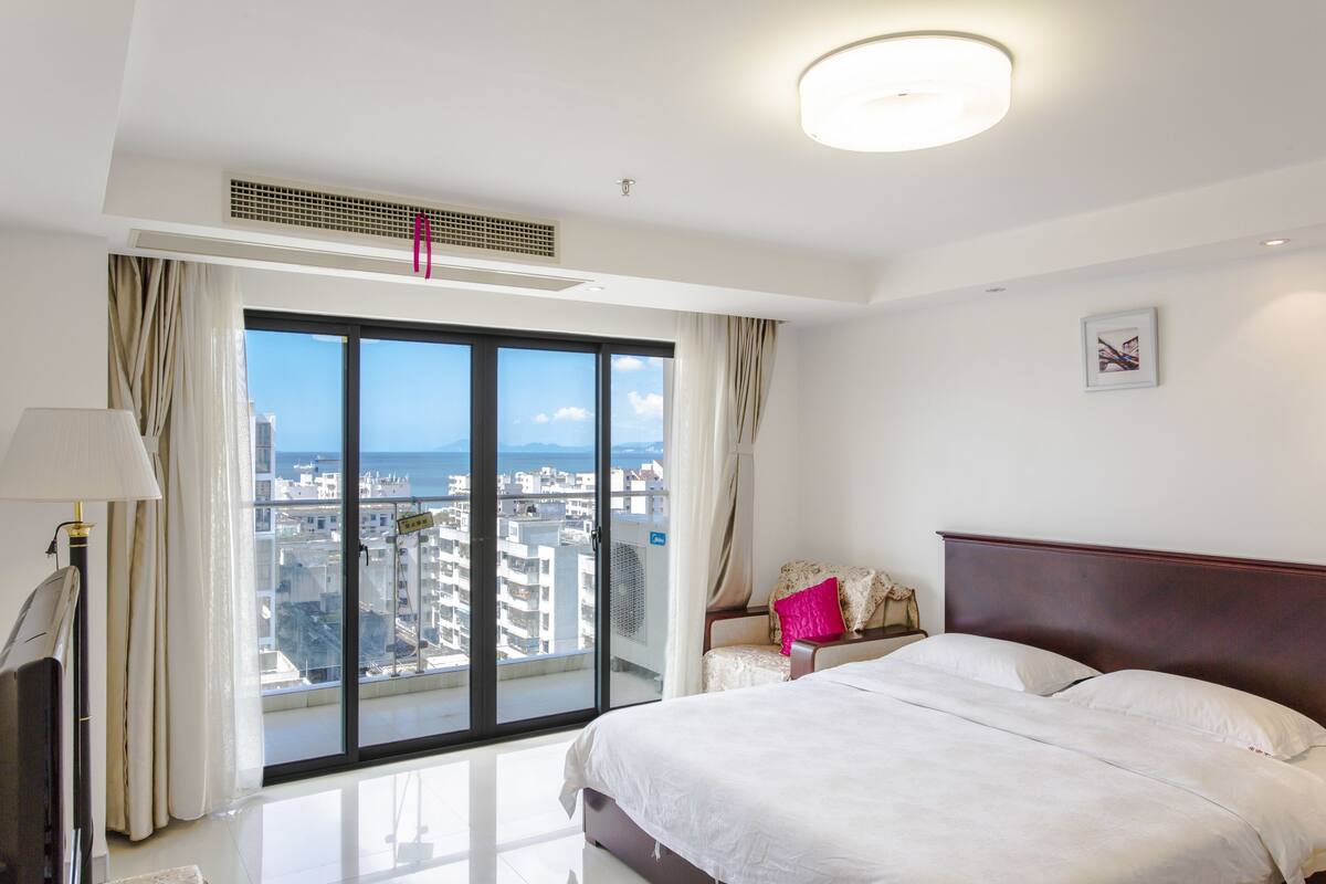 A hotel room with a large bed, a nightstand, a lamp, a window with curtains, and a view of the city.
