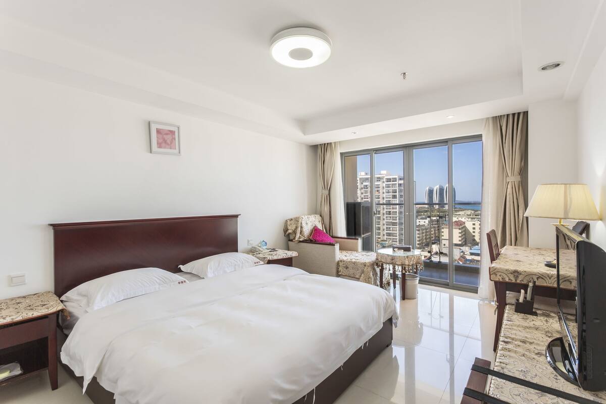 A hotel room with a large bed, a desk with a chair, a TV, and a view of the city through a large window.