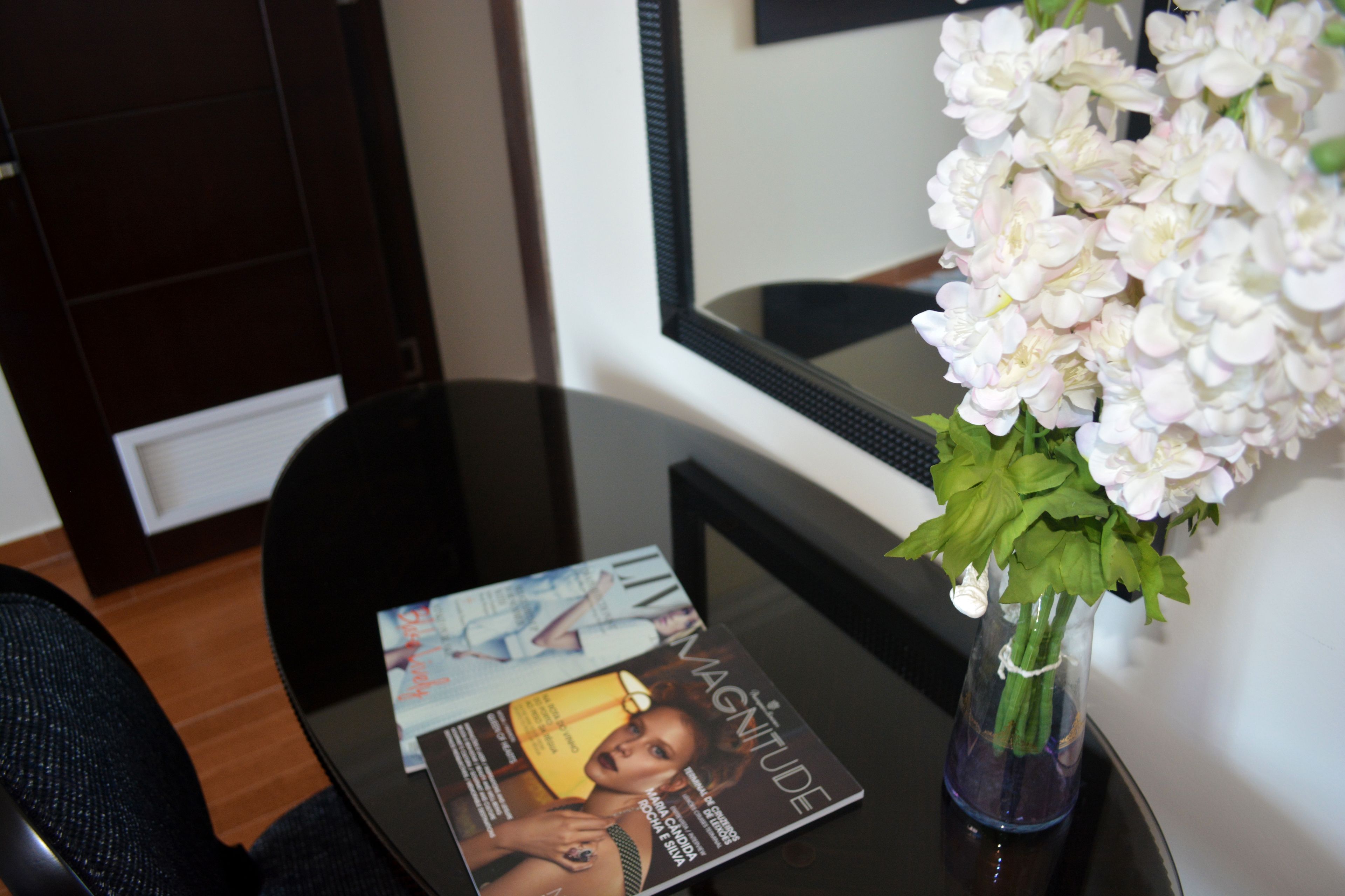 A magazine on a glass table, a vase with white flowers, and a dark wooden cabinet.