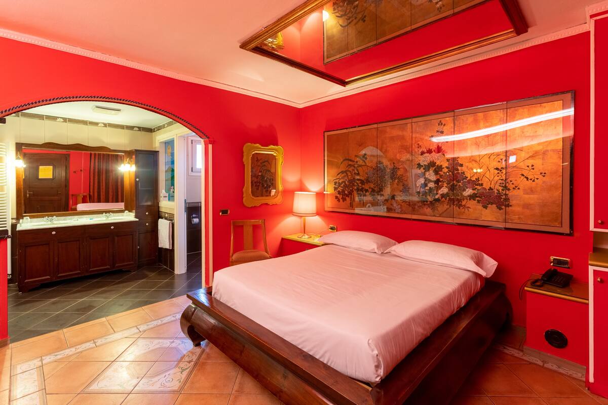 Une chambre dotée d’un grand lit, d’un miroir, d’une coiffeuse avec un lavabo et d’un grand tableau représentant des motifs floraux accroché au mur.