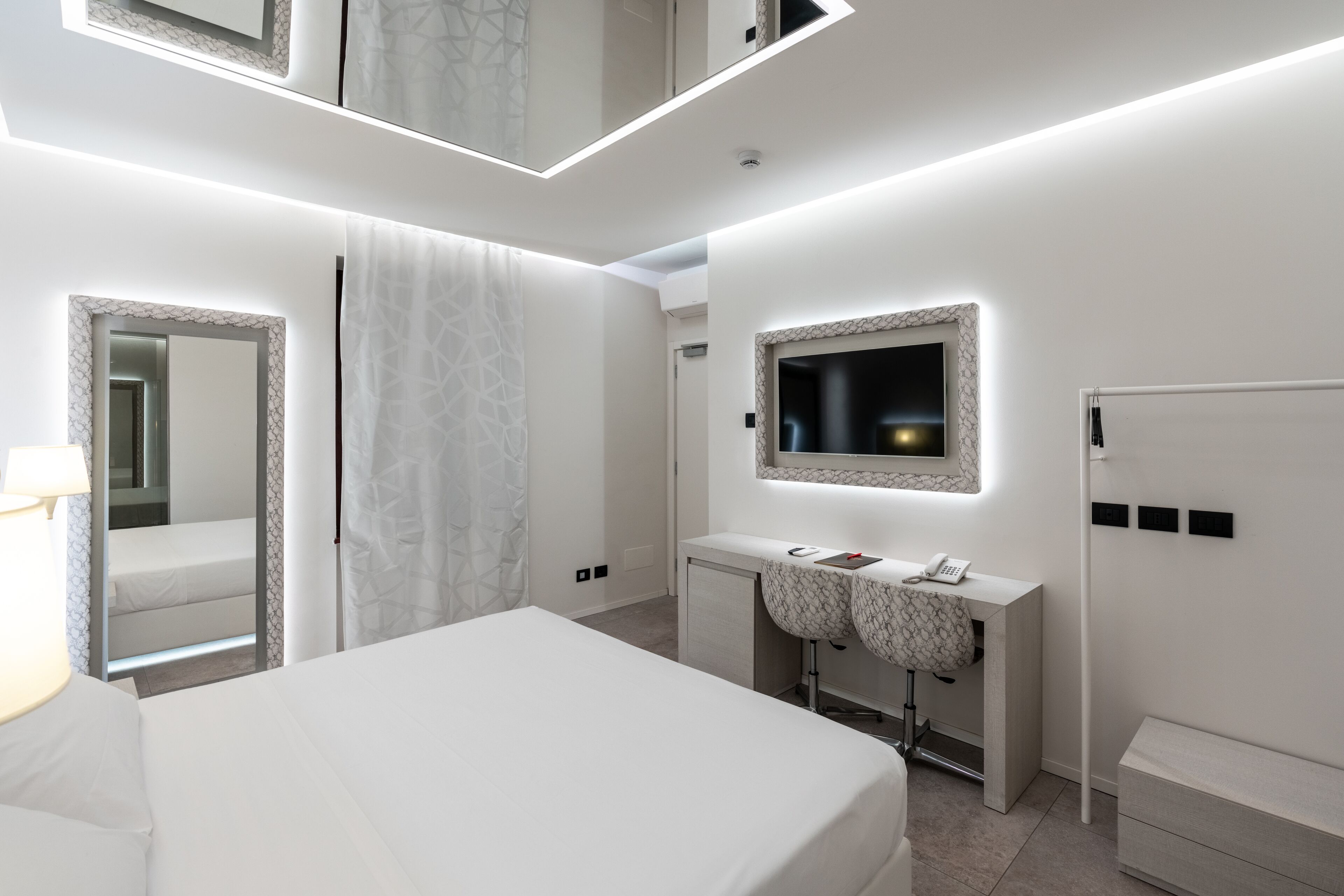A modern hotel room with a large bed, a vanity with a mirror, a desk, and a television.