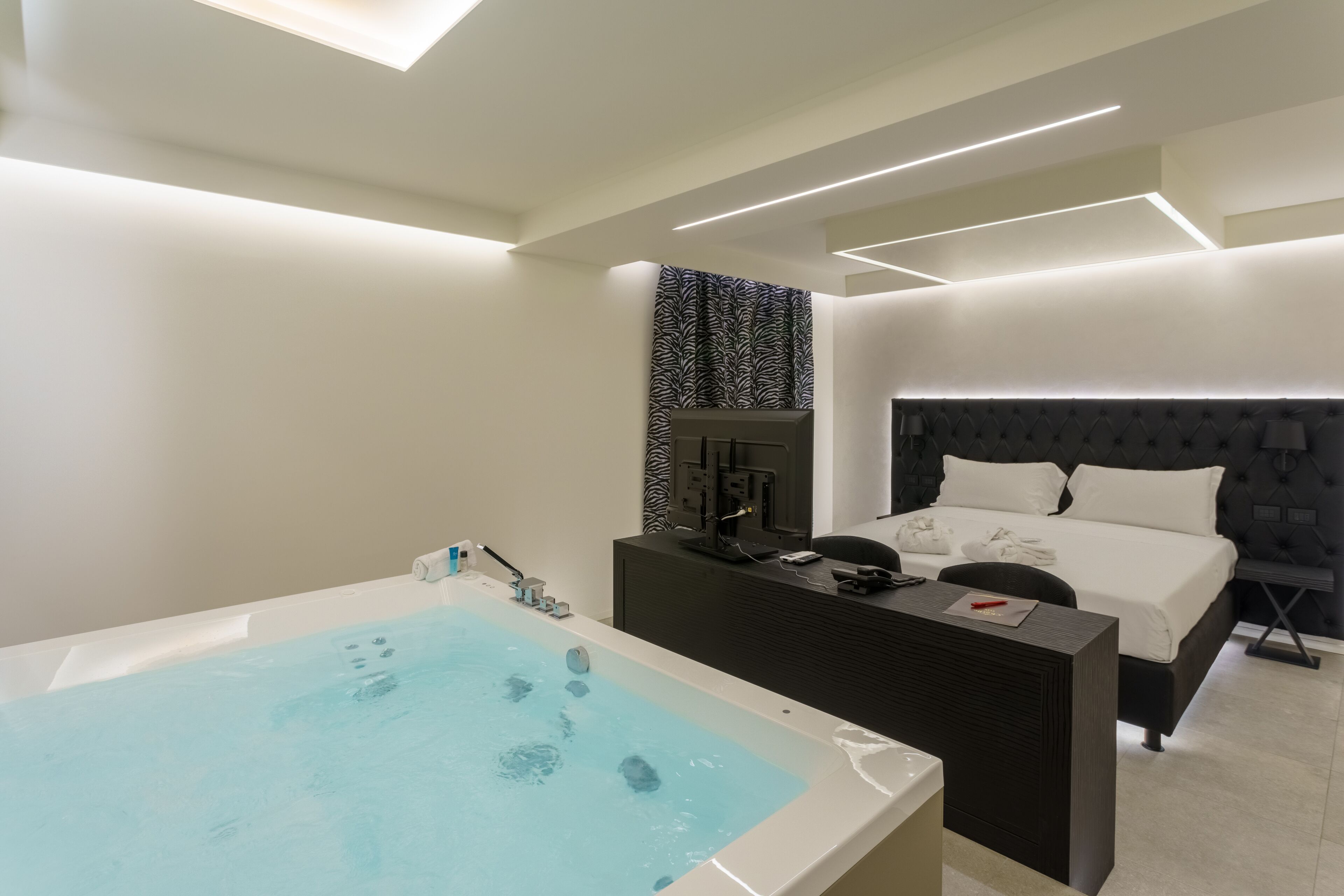 Una habitación de hotel moderna con una bañera grande, una cama con cabecera oscura y un escritorio pequeño con una computadora y accesorios.