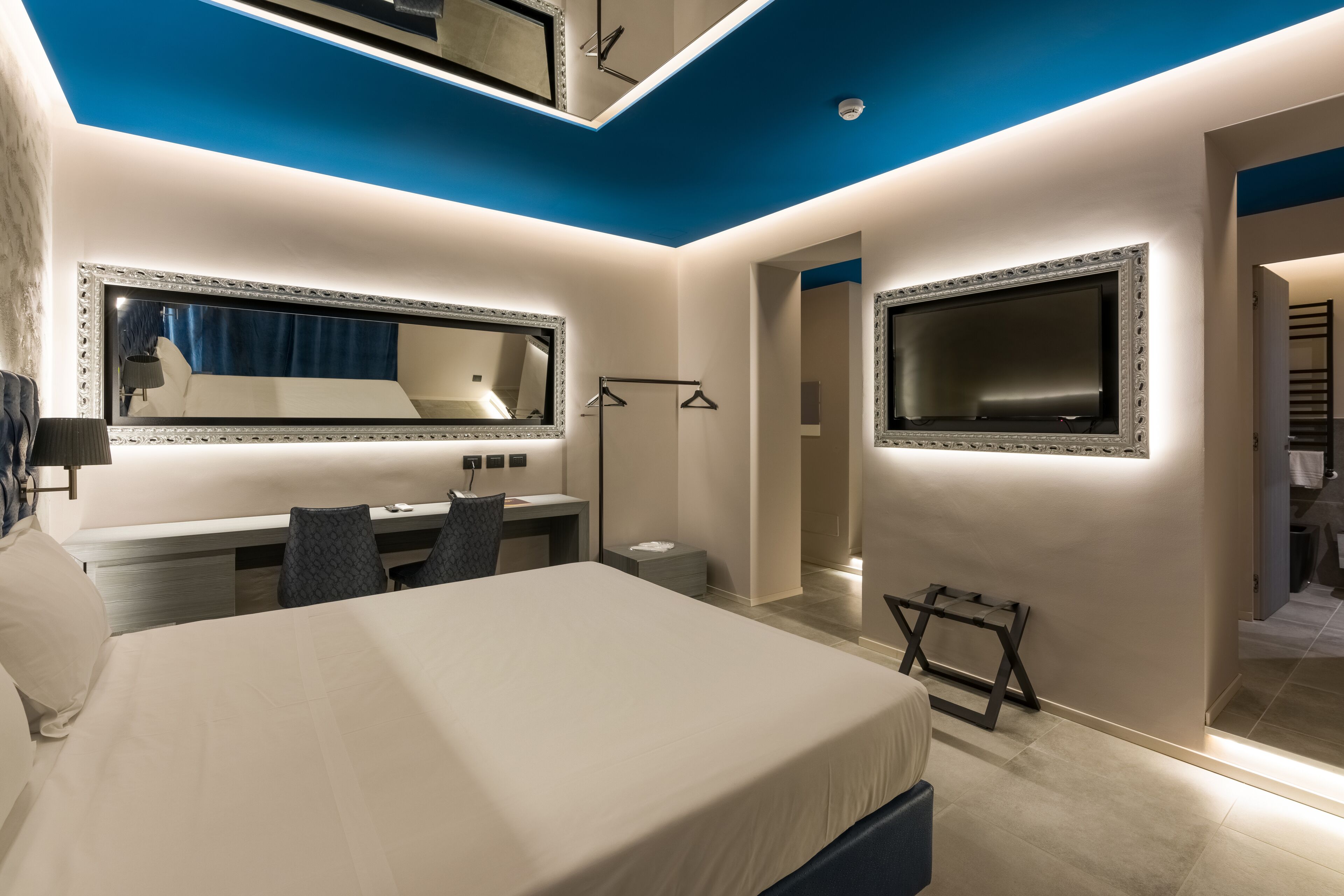 A modern hotel room with a large bed, a desk with a chair, a vanity with a mirror, and a TV mounted on the wall.