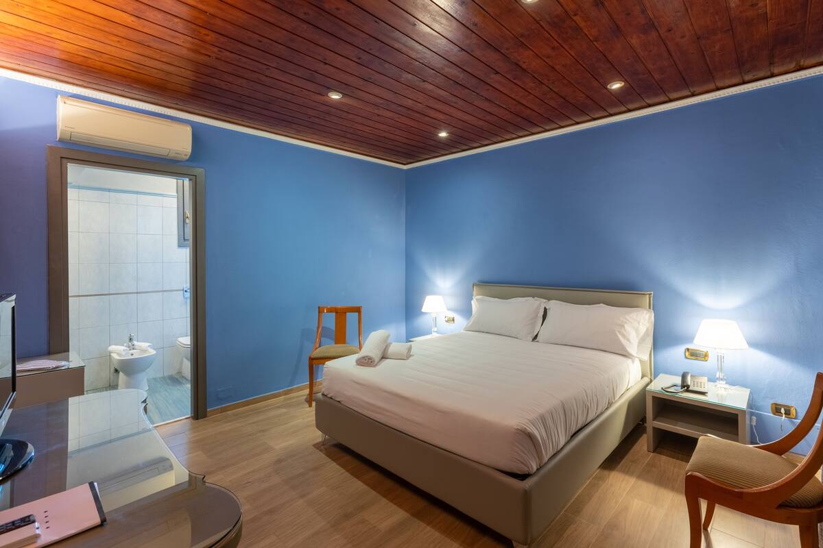 Une chambre d  ’hôtel avec un grand lit, des tables de chevet avec des lampes, une chaise, un bureau avec un ordinateur et une salle de bain visible à travers une porte en verre.