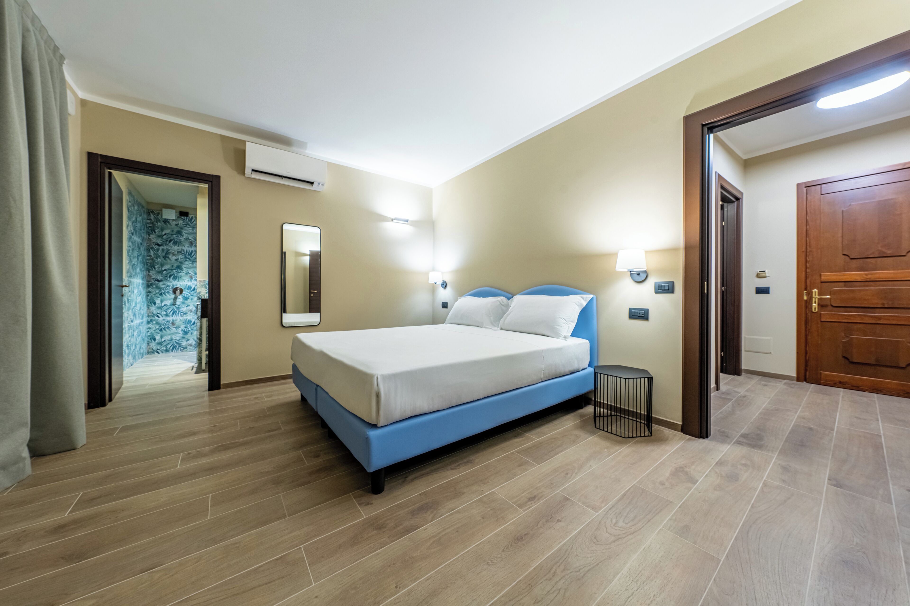 Habitación de hotel con una cama grande, piso de madera, baño con ducha azul y puerta de madera.