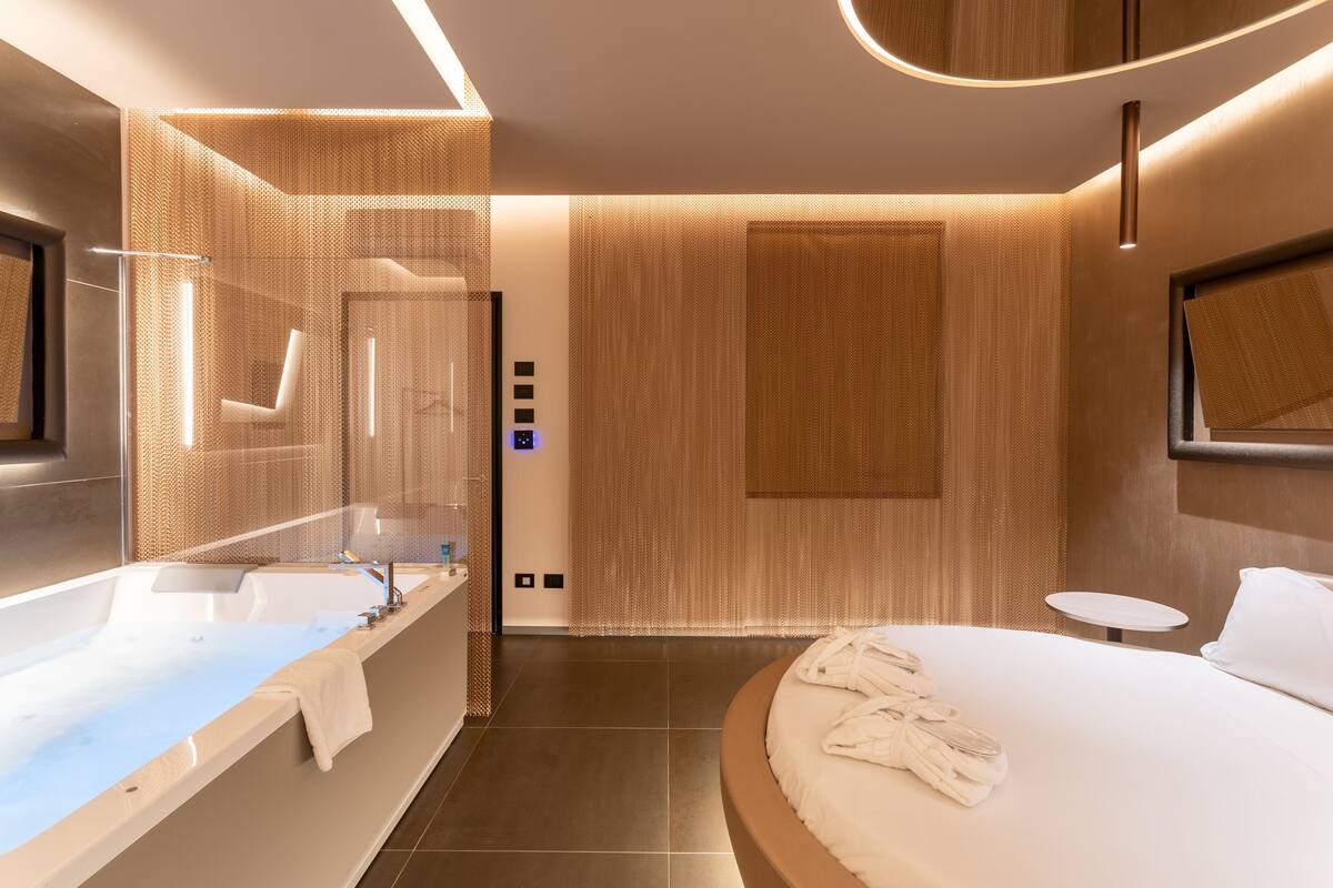 Une chambre d’hôtel moderne dotée d’une grande baignoire, d’un espace douche séparé, d’un lit rond avec des serviettes et d’une petite table.
