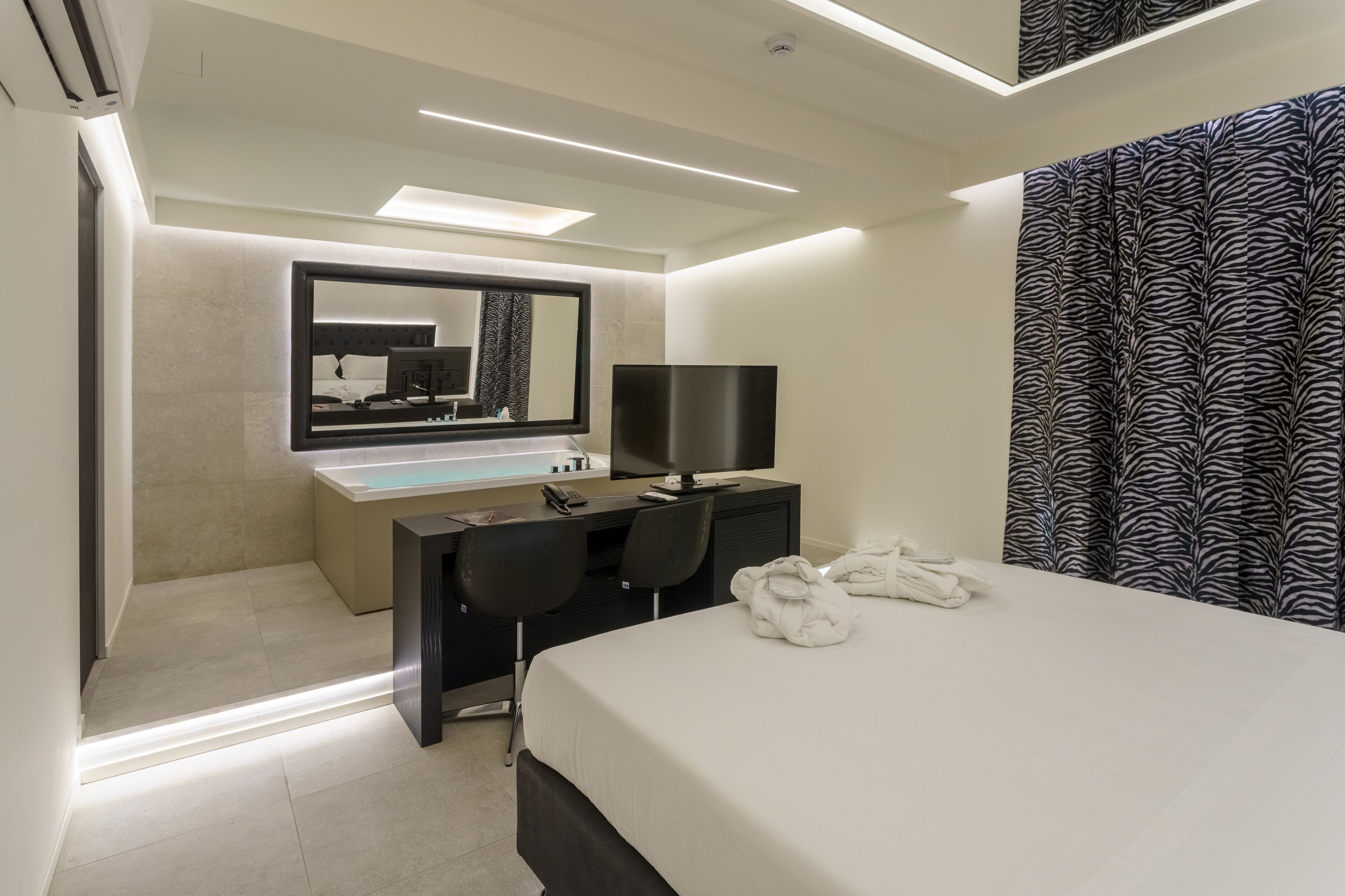 Ein modernes Hotelzimmer mit einem Bett, einem Schreibtisch mit Computer, einem Spiegel und einem Badezimmer.