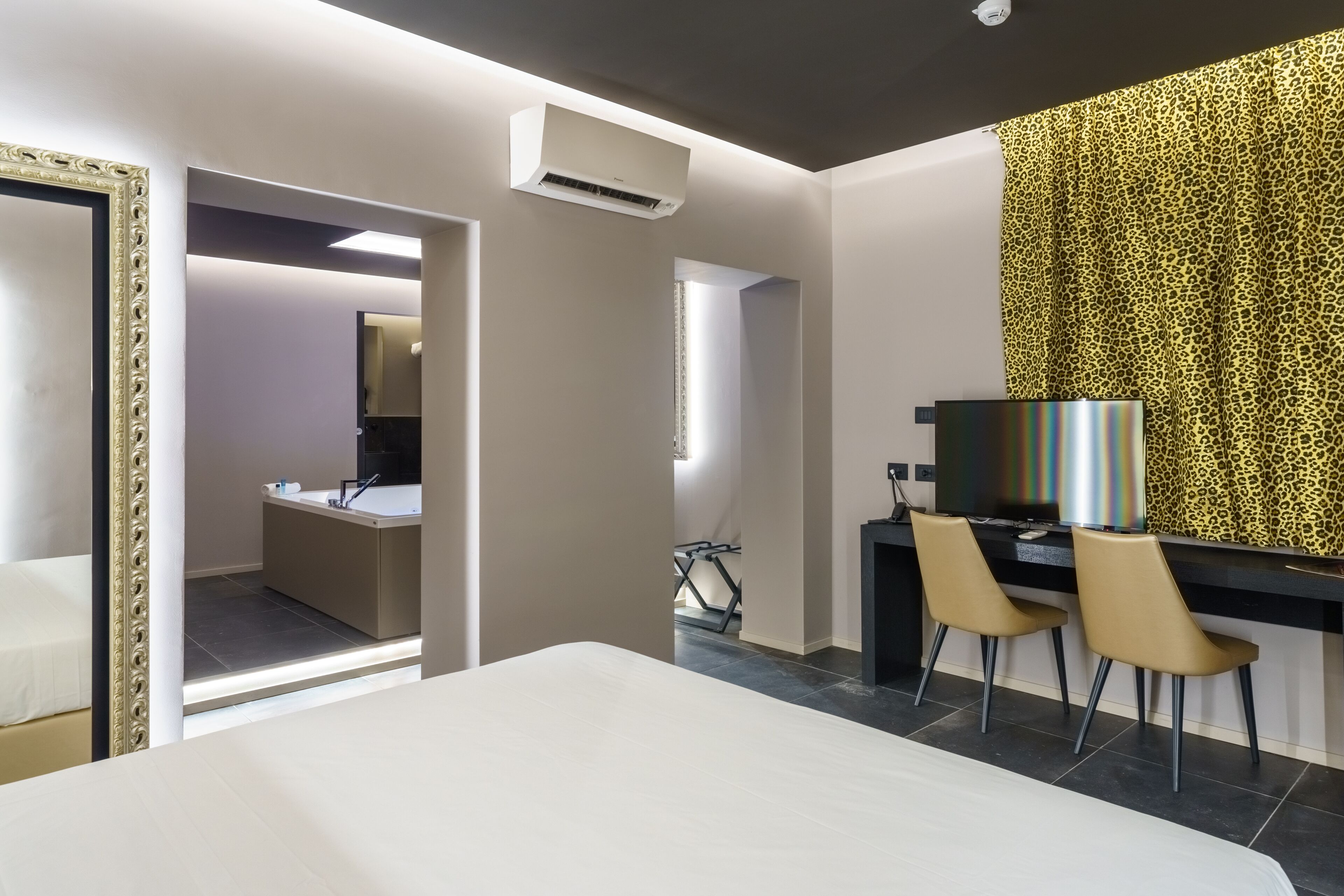 Ein Hotelzimmer mit einem Bett, einem Schreibtisch mit zwei Stühlen, einem Fernseher und einem Spiegel. Ein Badezimmer ist durch eine offene Tür sichtbar.