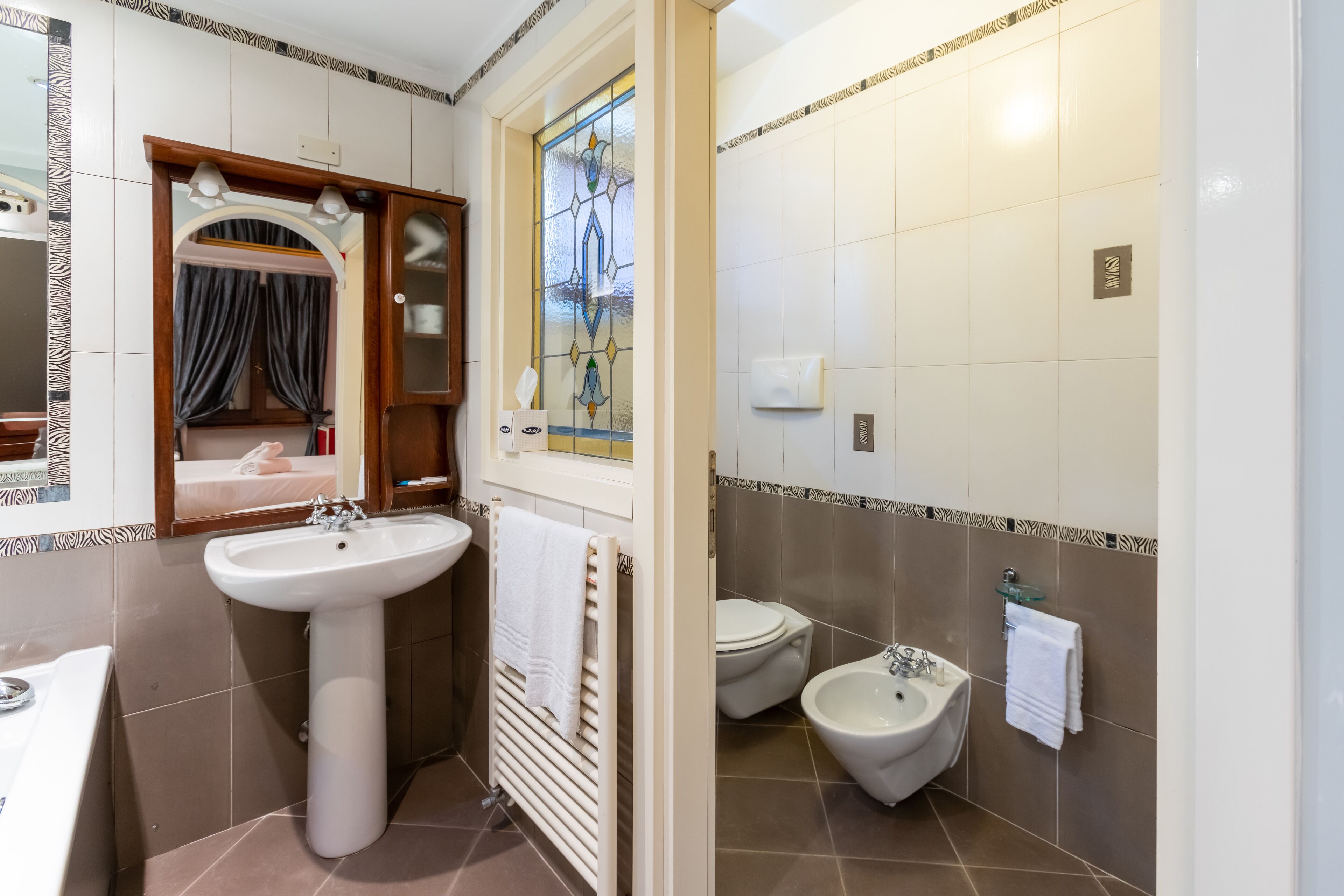 Ein Badezimmer mit einem Waschbecken auf Säule, einem Bidet, einem Spiegel und einem Fenster mit Buntglas.