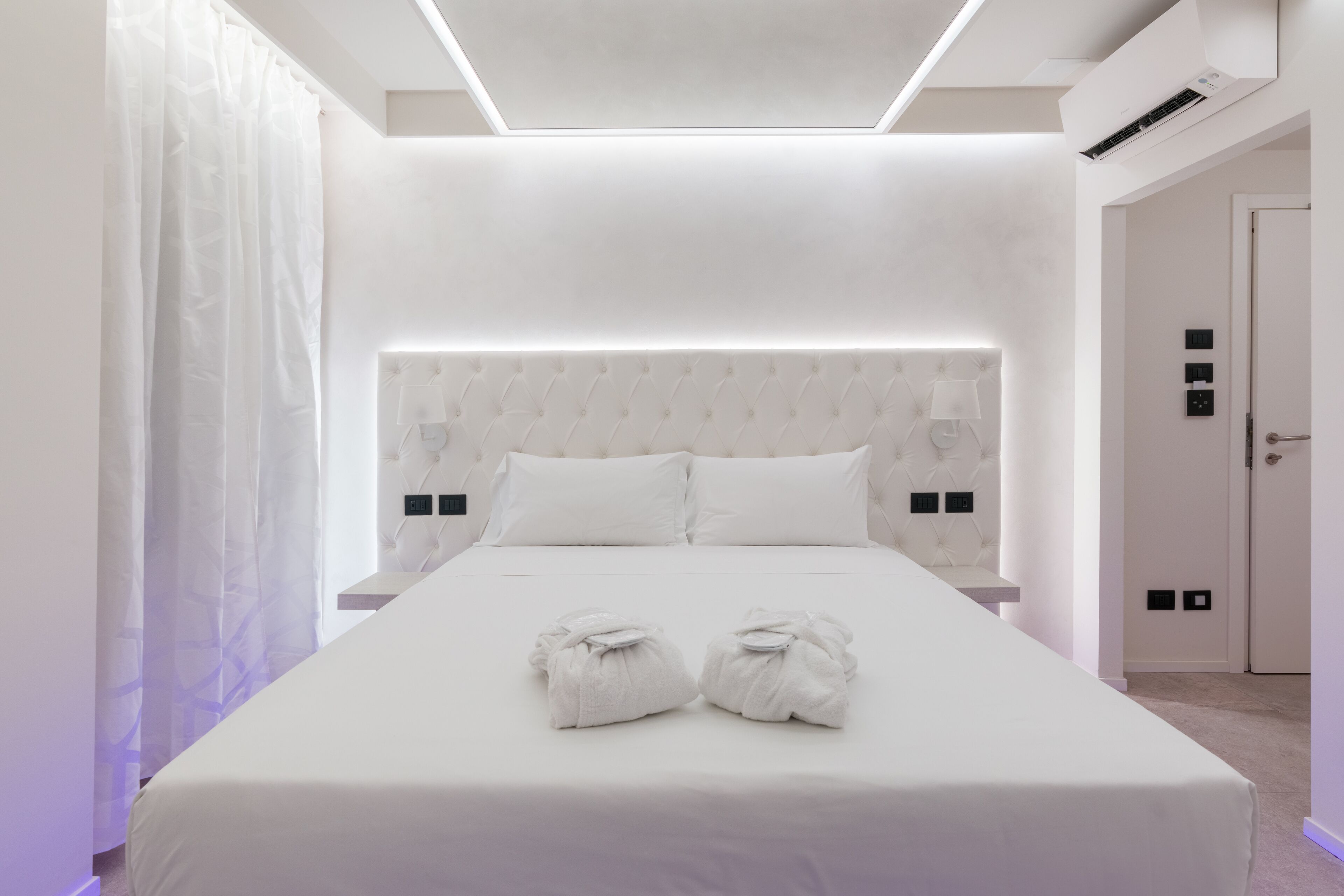 Camera d'albergo moderna con un grande letto, lenzuola bianche, testiera imbottita e due asciugamani sul letto.