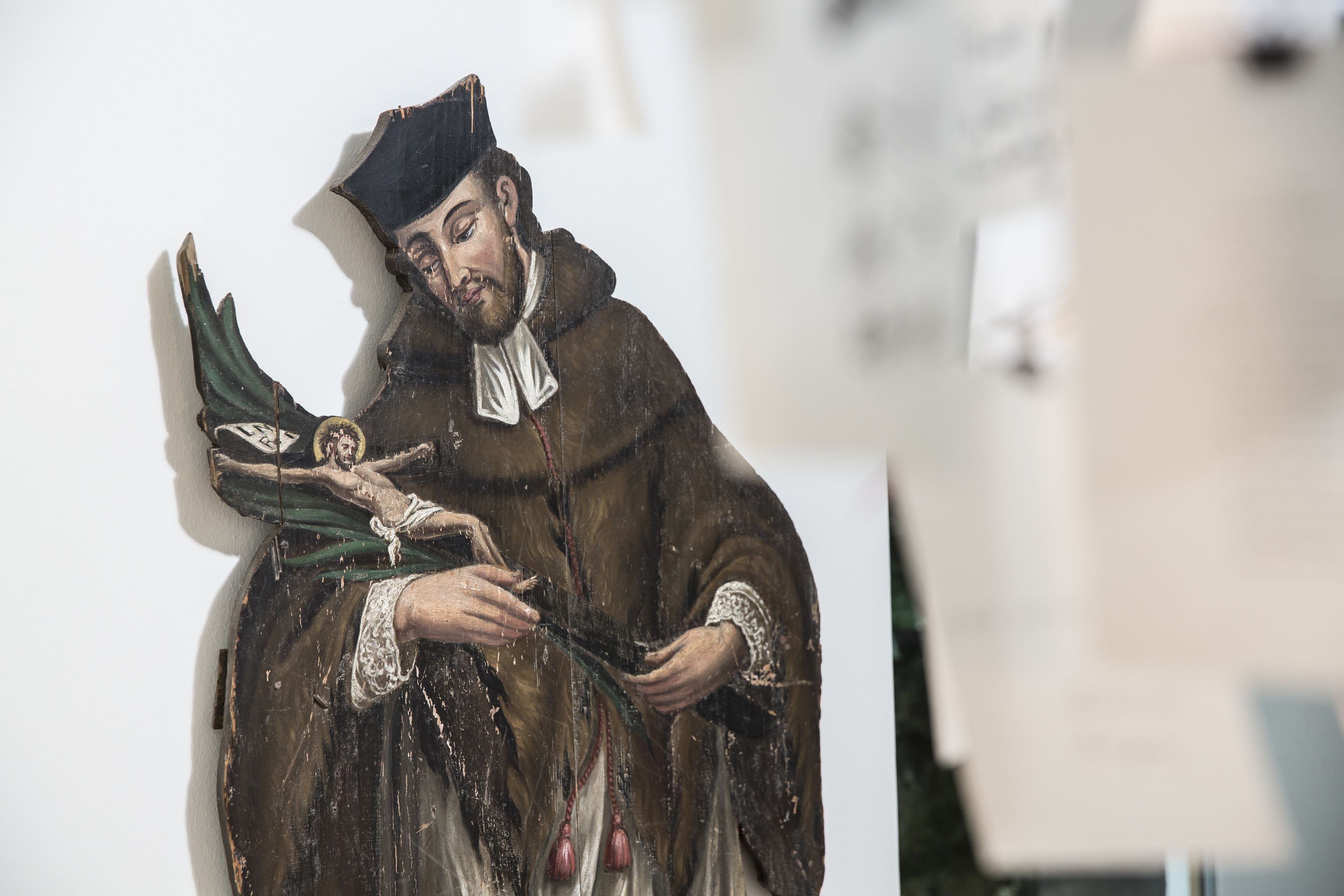 A statue of a robed figure holding a book and a staff, with a background of a white wall and a partially visible poster.