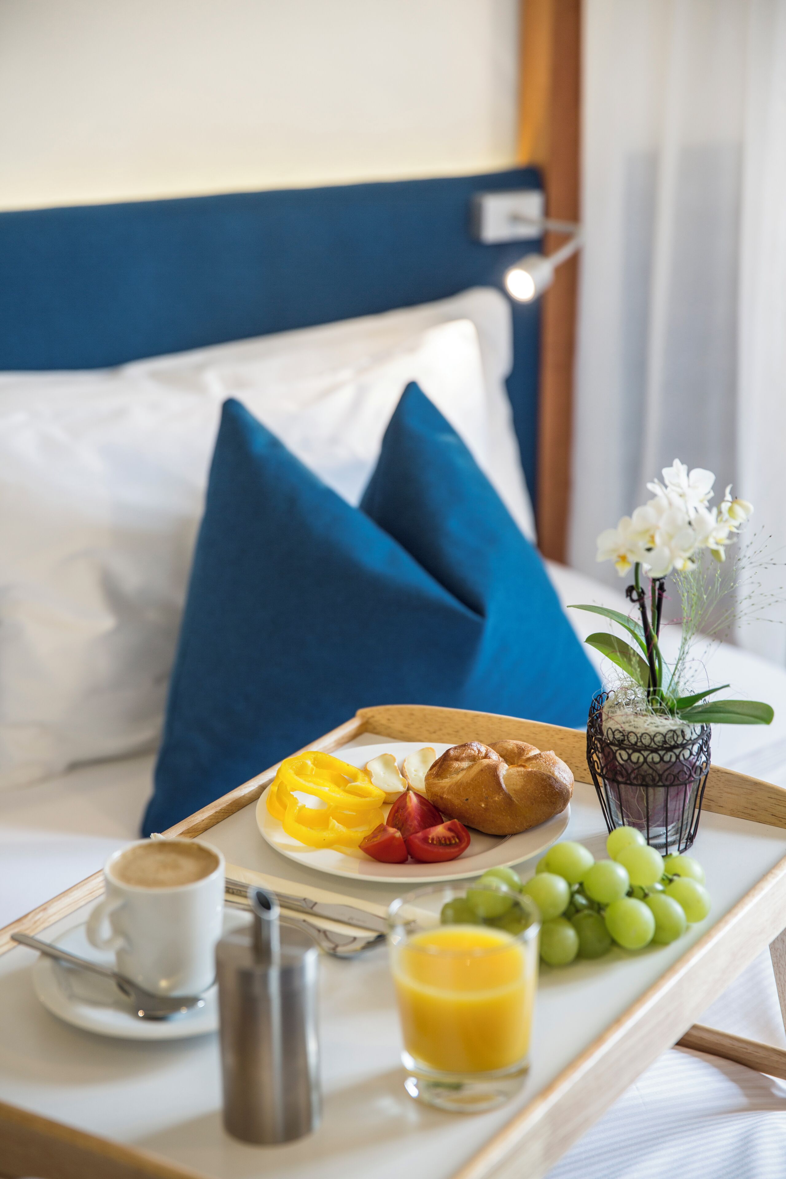 Un plateau de petit-déjeuner avec des croissants, des fruits et du jus, posé sur un lit avec des oreillers bleus.