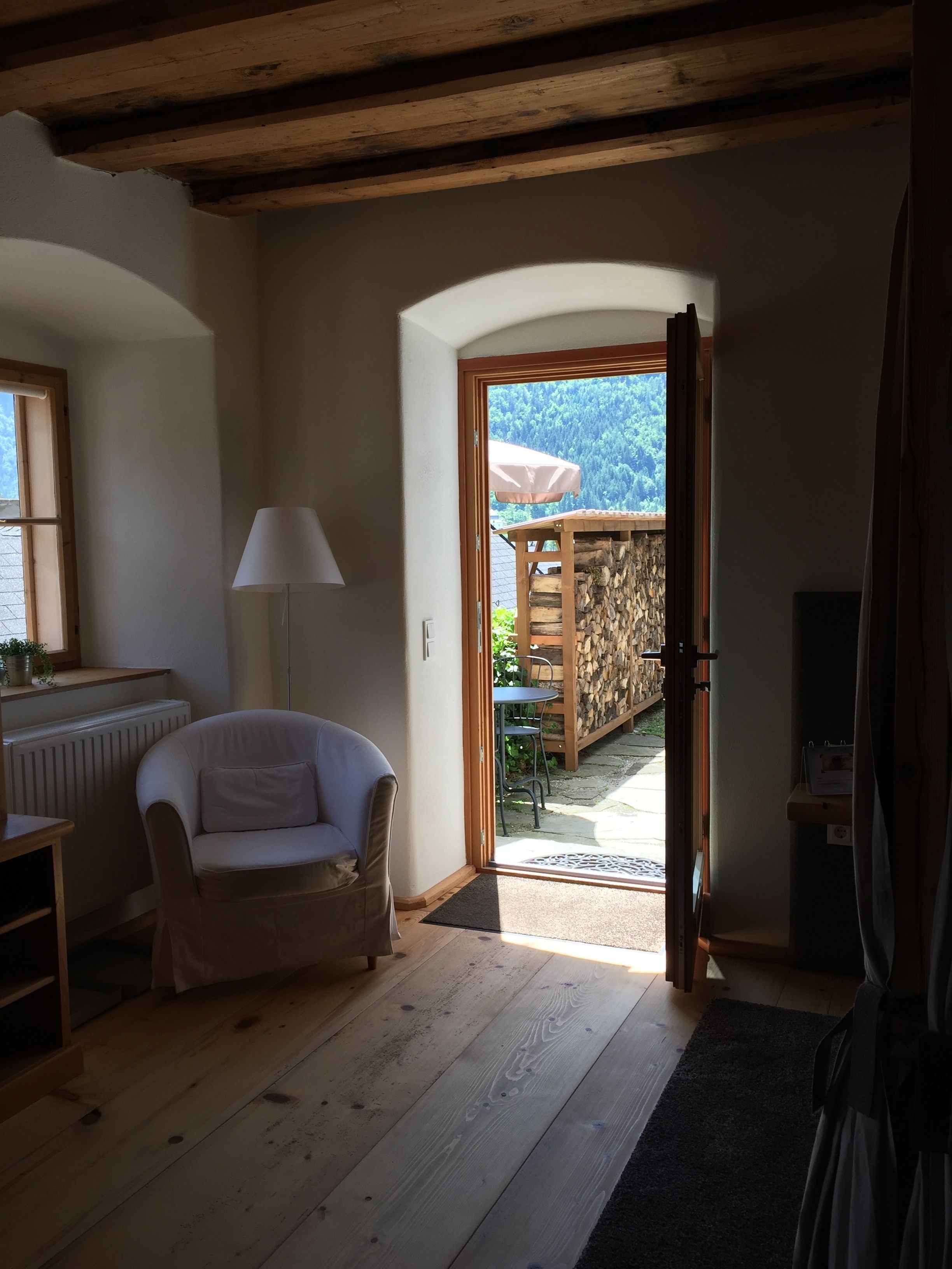 A cozy room with a white armchair, a lamp, and a view of a wooden deck and mountains through an open door.