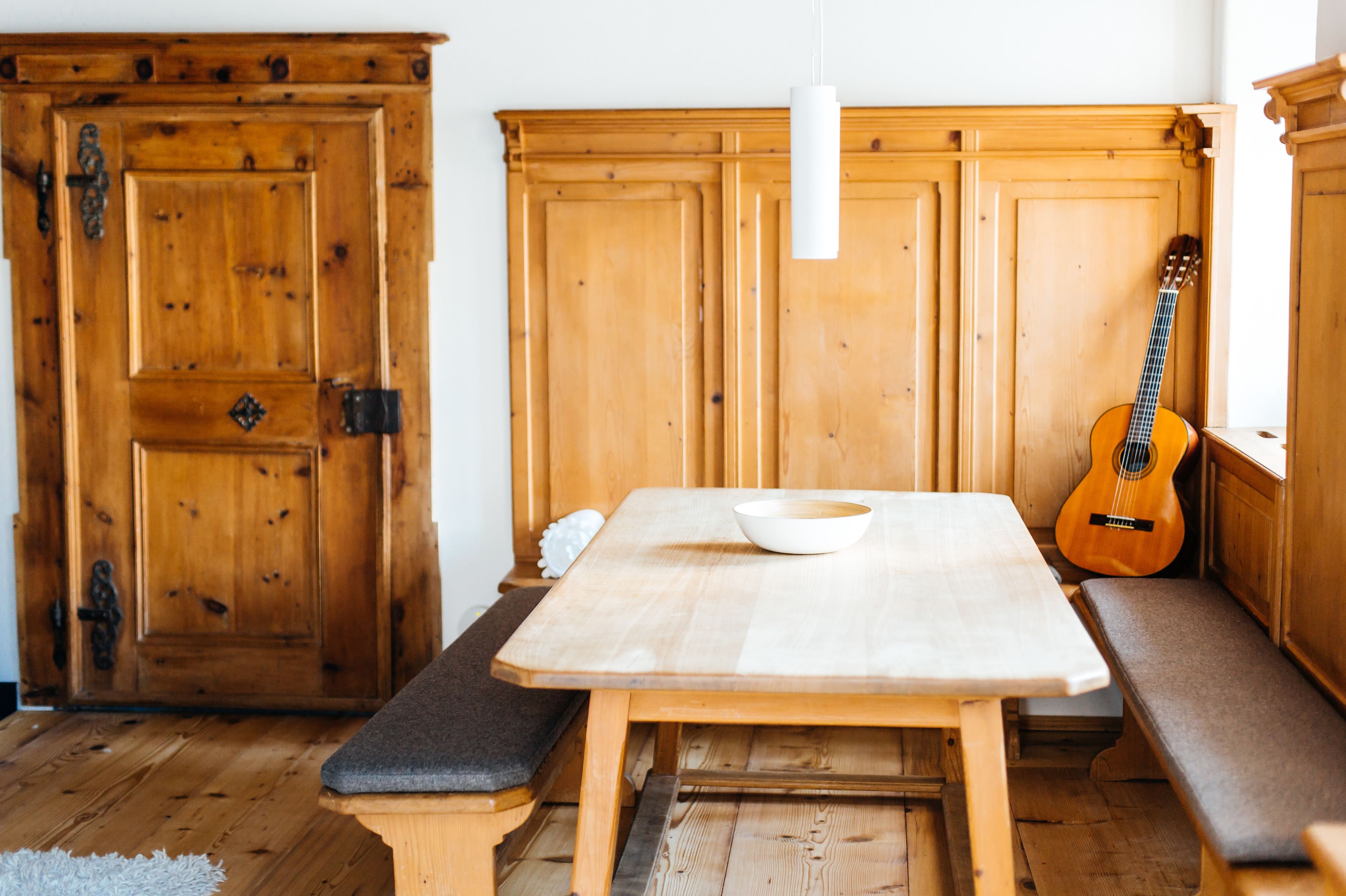 A wooden dining table with a bowl, benches, and a guitar leaning against a wooden cabinet.