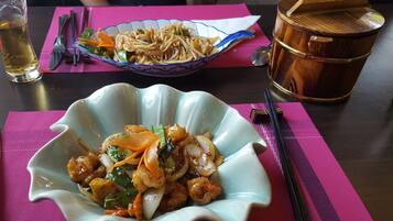 Un tazón de vegetales salteados y fideos sobre una mesa, con palillos chinos, una jarra de cerveza y un recipiente de madera tradicional.