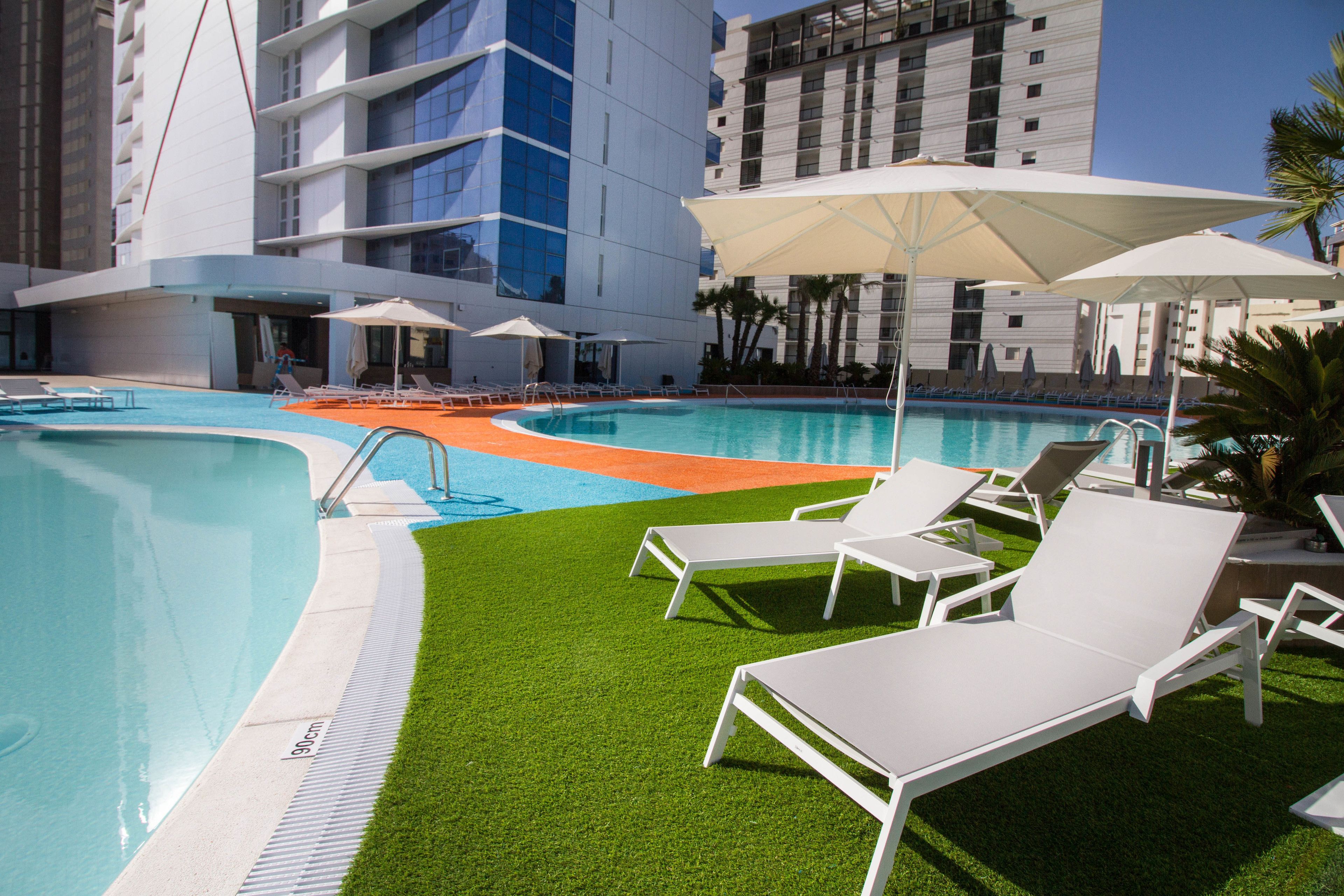 Piscina com espreguiçadeiras brancas e guarda-sóis, cercada por gramado e um edifício com várias andares e varandas.
