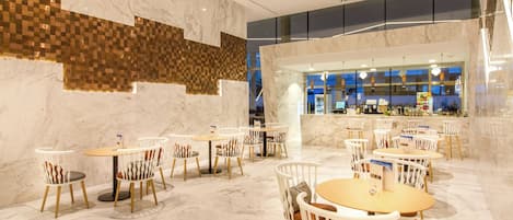 A spacious indoor area with marble flooring and modern furniture, including round tables and chairs, and a visible bar area in the background.
